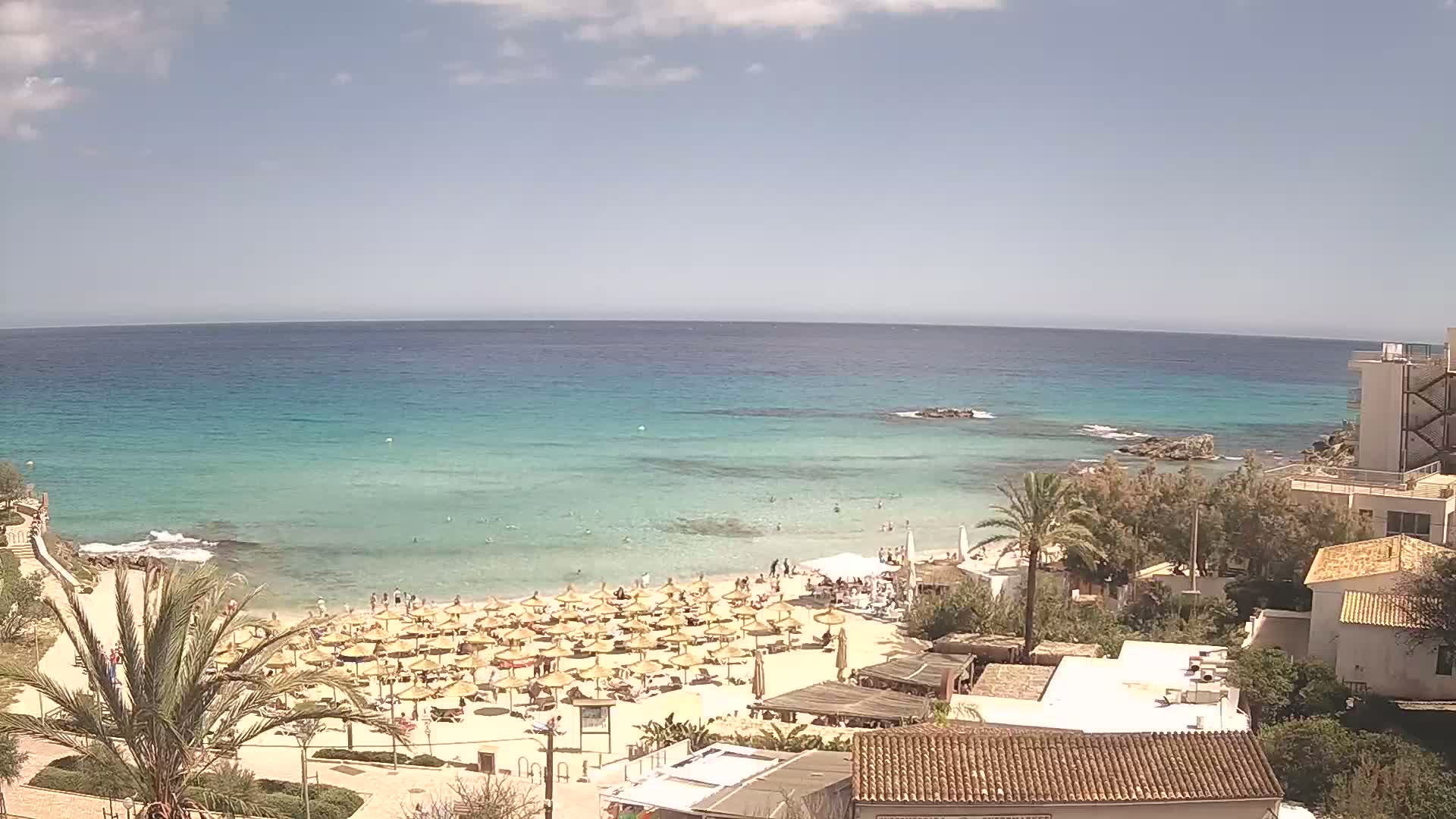 A beach with many straw umbrellas and sunbathers is situated next to a calm, turquoise sea under a mostly sunny sky.