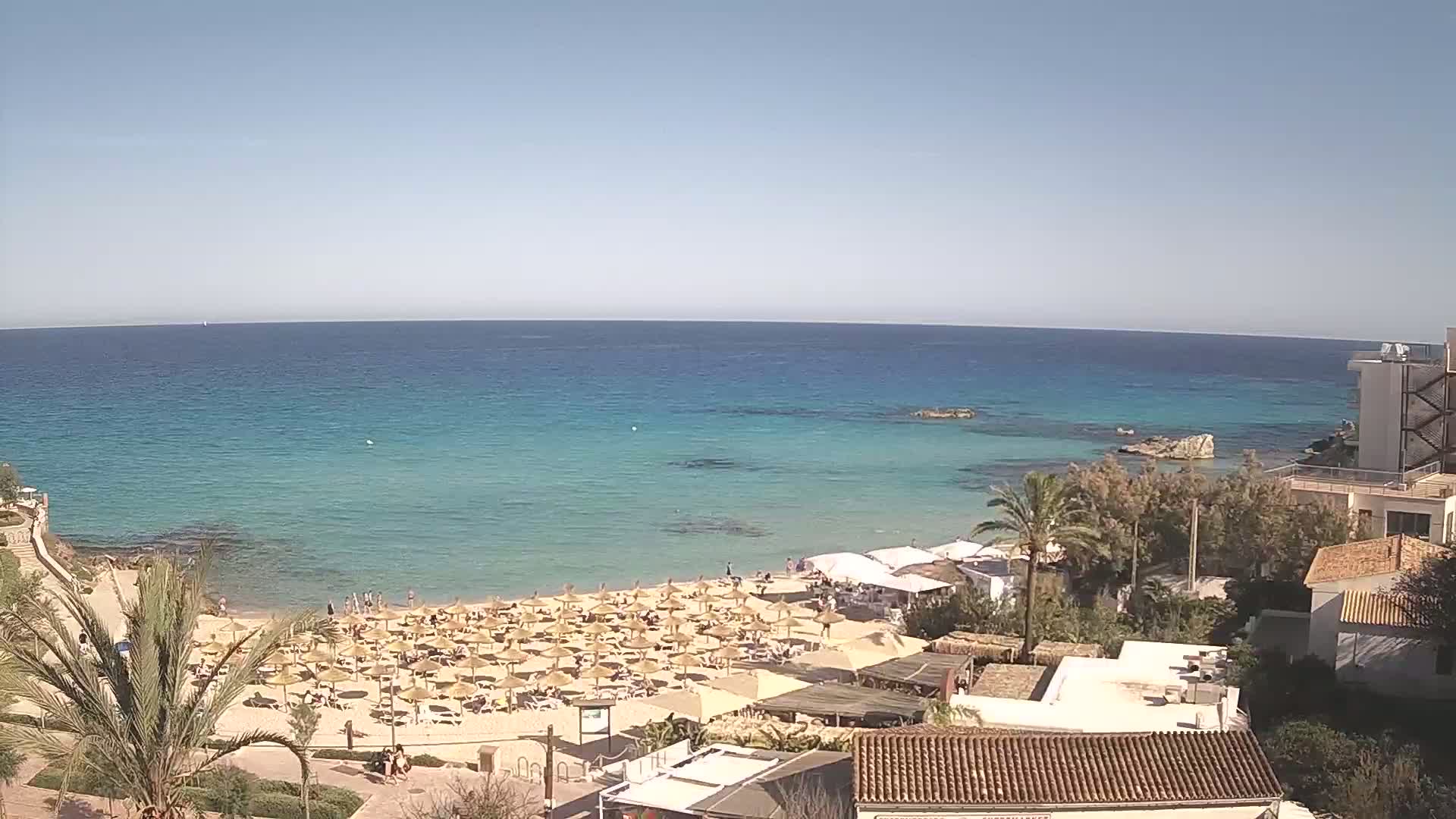 A sandy beach with numerous straw umbrellas is situated next to a calm, clear turquoise sea under a bright sunny sky, backed by buildings and vegetation.