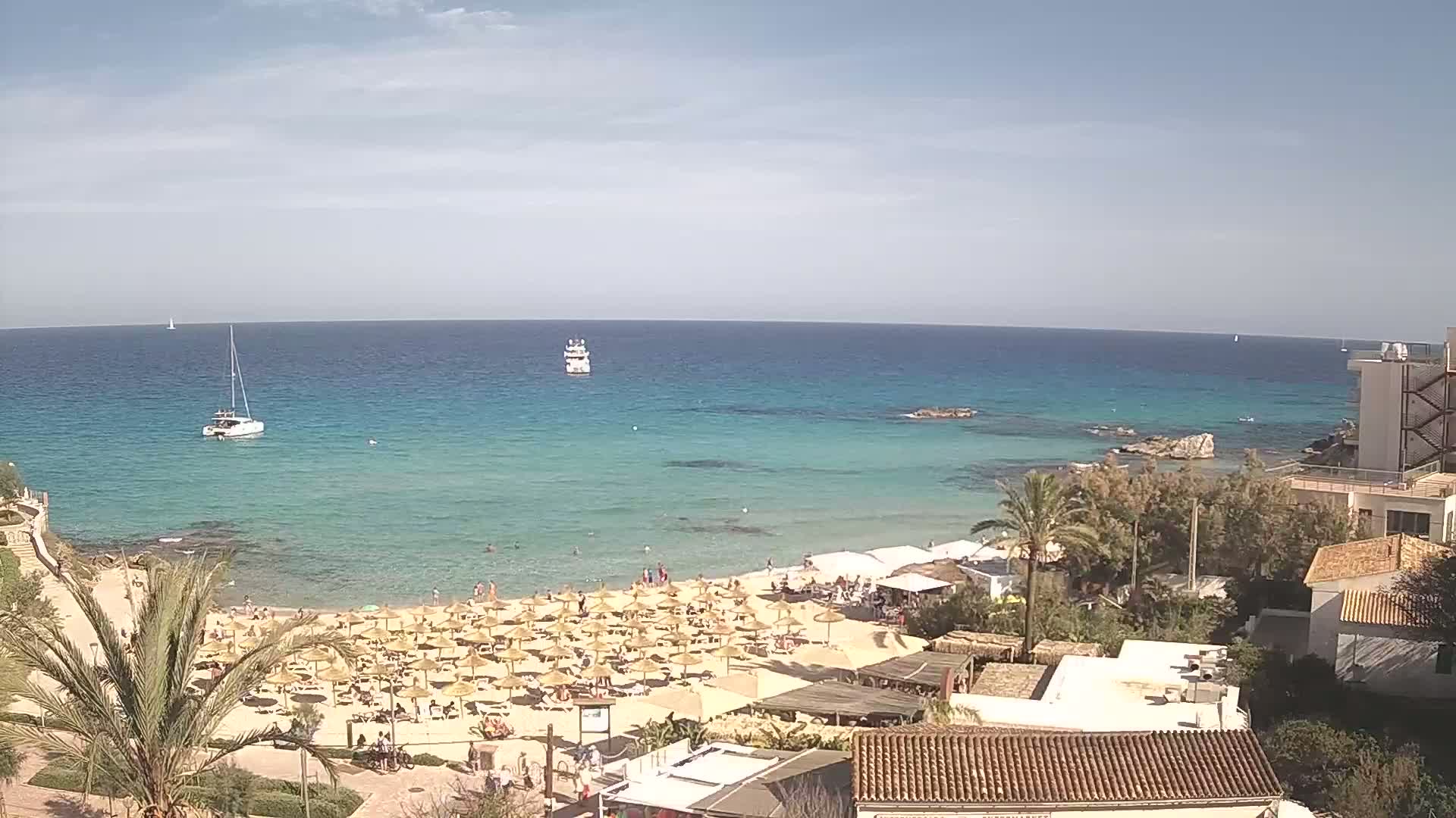 A sandy beach with numerous straw umbrellas and sunbathers is situated beside a calm, turquoise sea, under a partly cloudy sky; several boats are visible offshore.