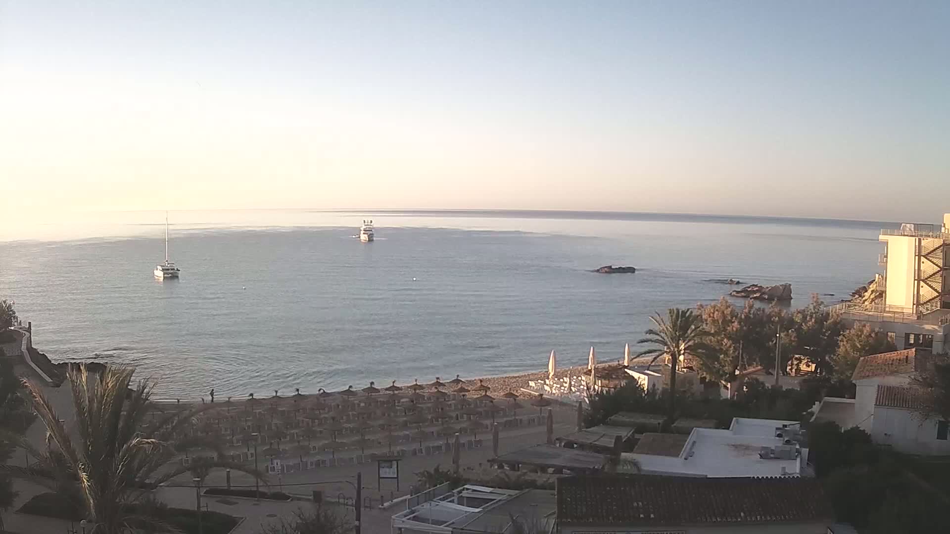 A calm, clear day shows a sandy beach with rows of umbrellas, palm trees, buildings, and two boats on a tranquil sea.