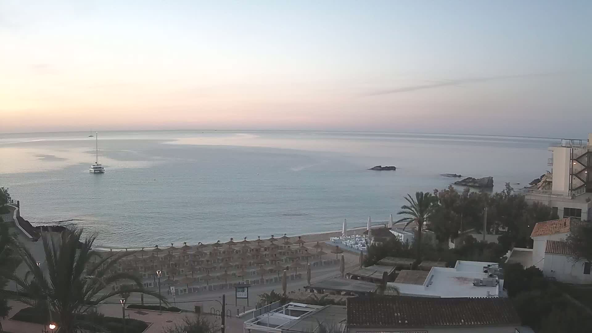 A calm, pale-pink sunrise illuminates a beach with rows of thatched umbrellas, a small boat on the water, and buildings along the shore.