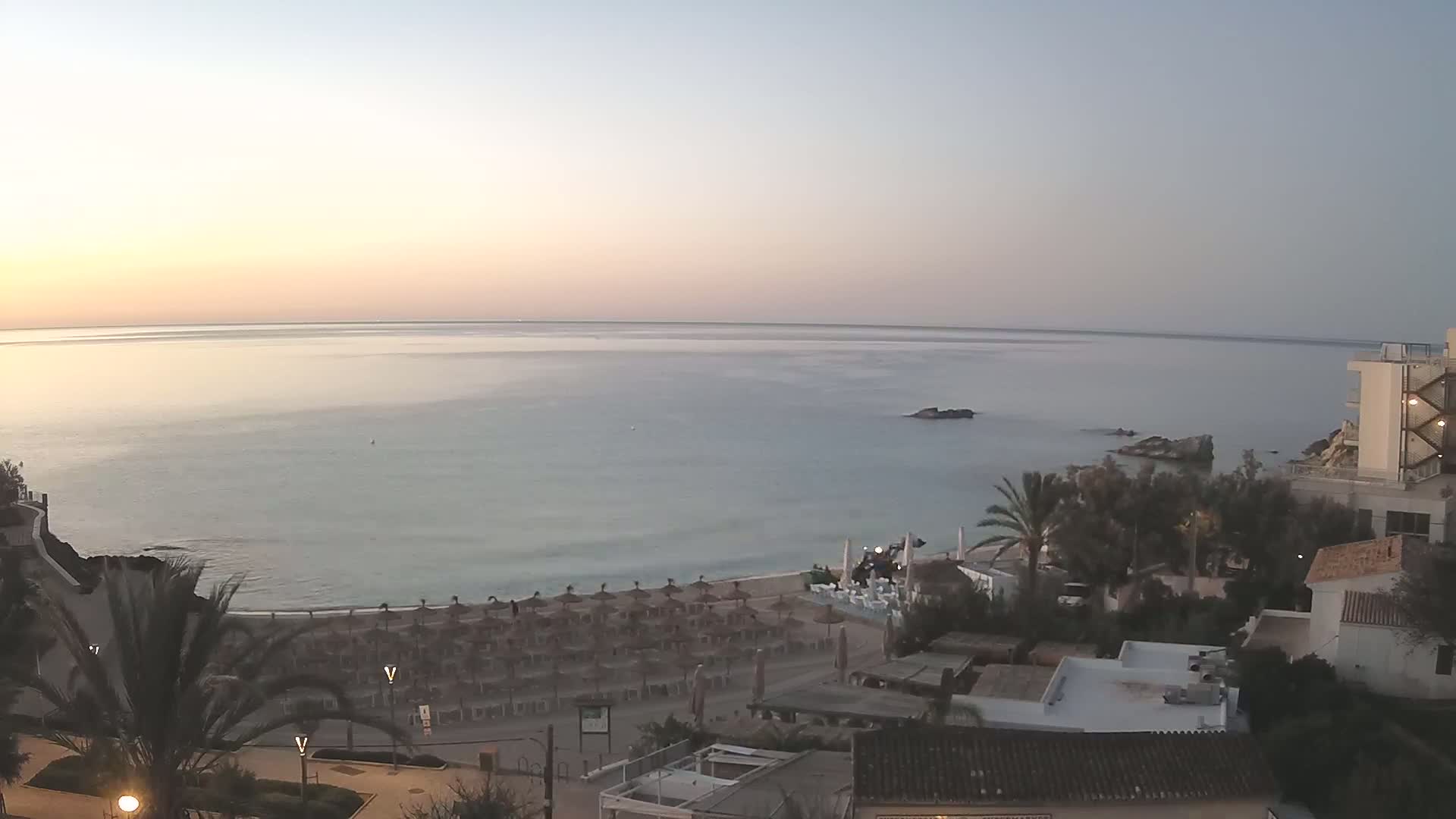 A calm, pale-blue sea at sunrise is seen from an elevated vantage point, overlooking a sandy beach with numerous closed umbrellas and several buildings nestled among palm trees.