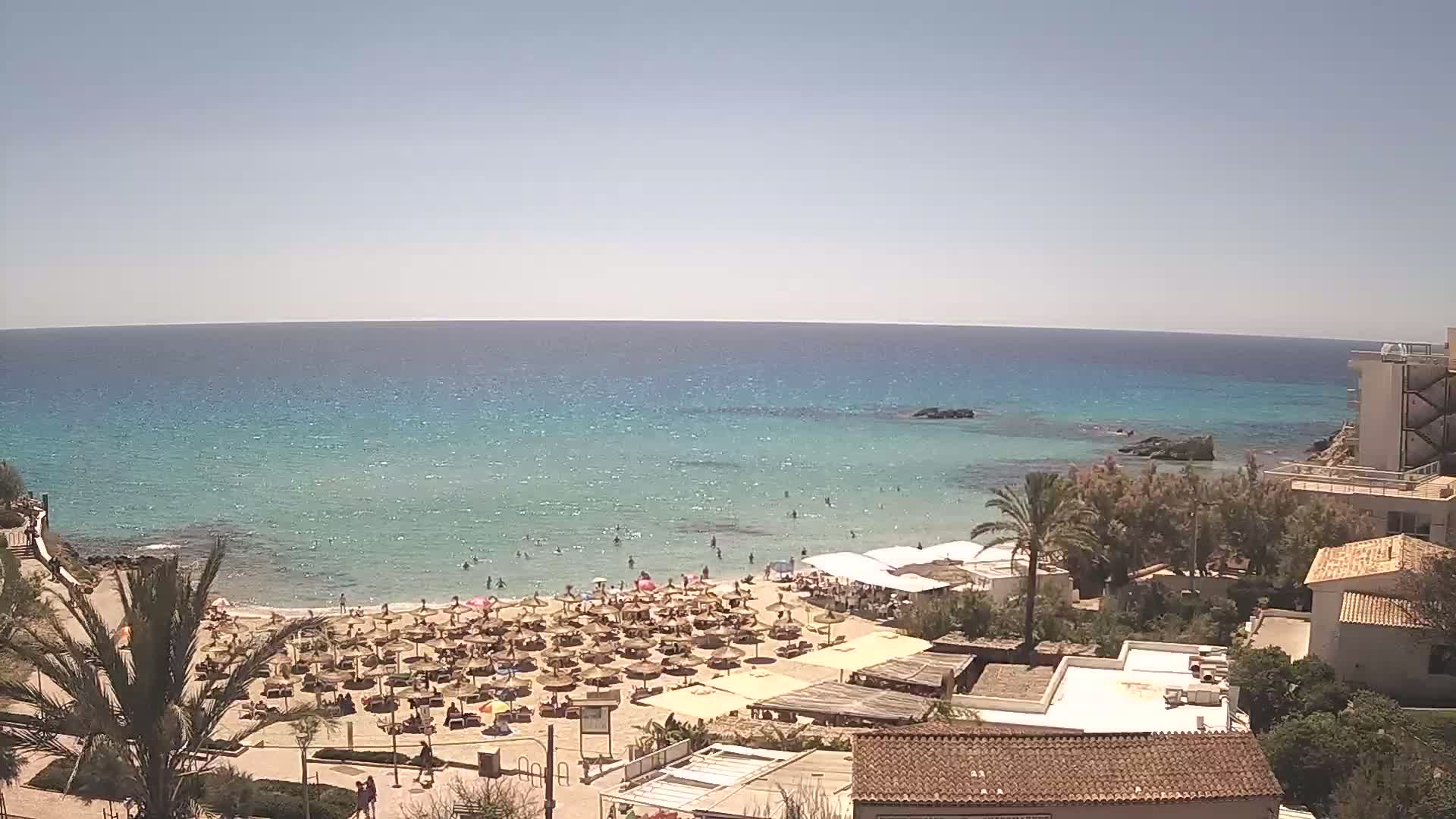 A sandy beach with numerous umbrellas and sunbathers is situated next to a calm, turquoise sea under a clear blue sky.