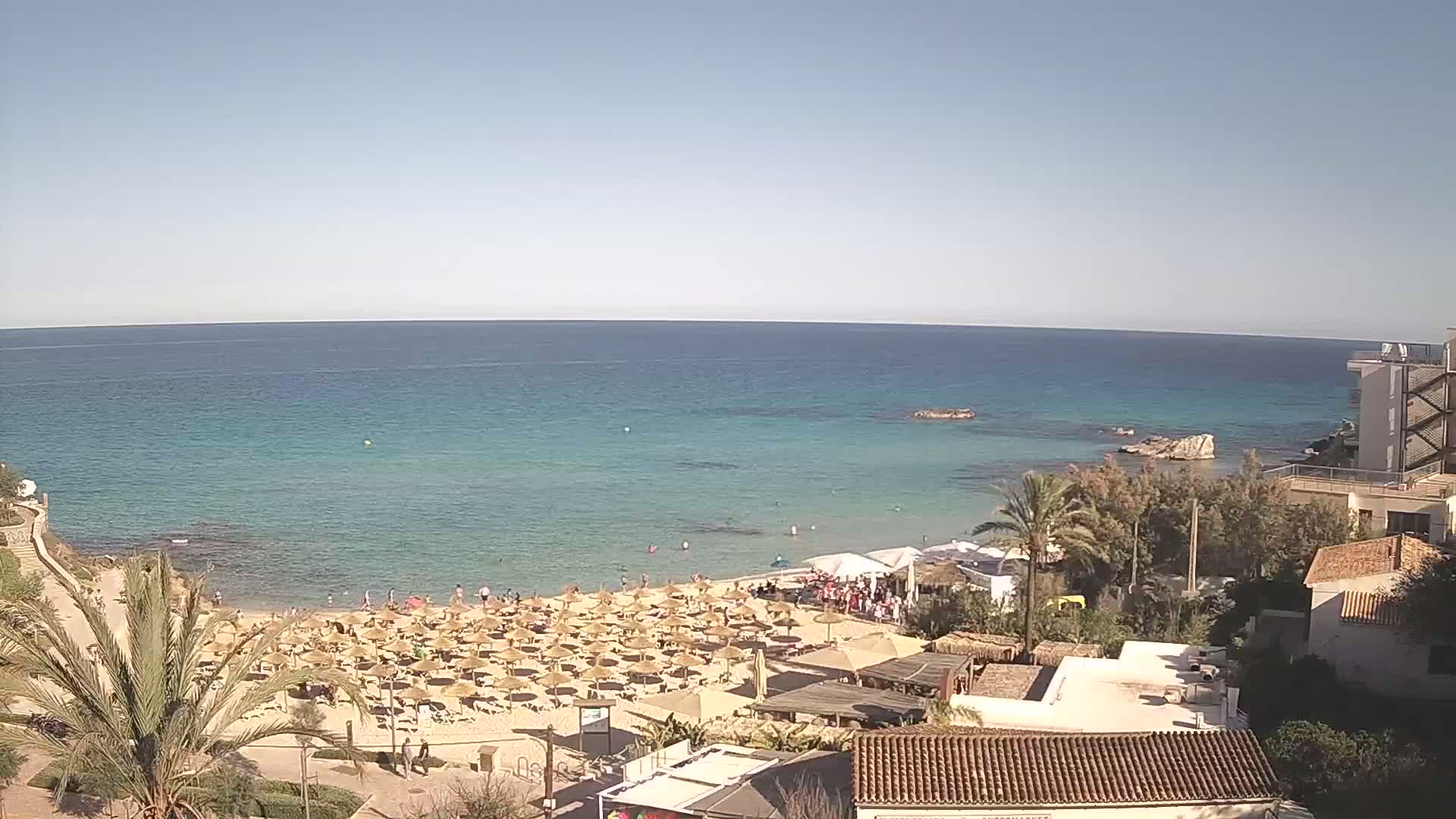 A sandy beach with numerous umbrellas and people is situated next to a calm, clear turquoise sea under a bright, sunny sky.