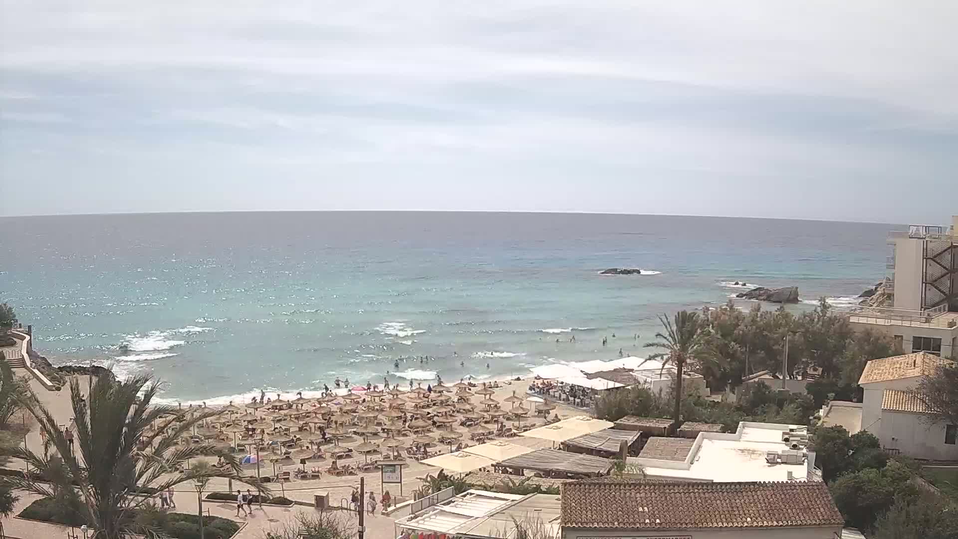 A sandy beach with numerous umbrellas and beachgoers is situated next to a calm, light-blue ocean under a mostly cloudy sky.