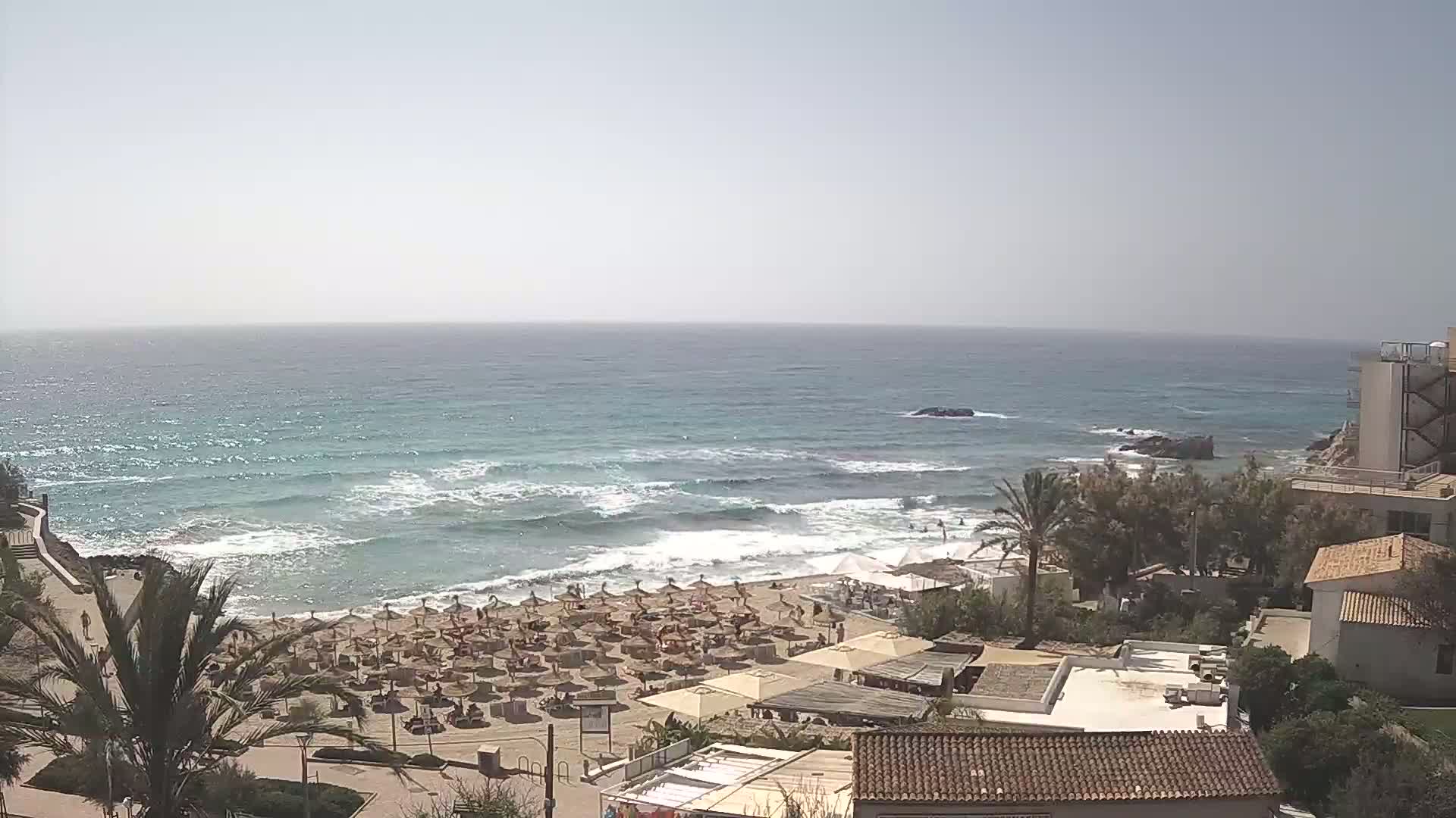 A sandy beach with numerous umbrellas and people is situated next to a calm sea with small waves under a bright, sunny sky, overlooking a collection of buildings.