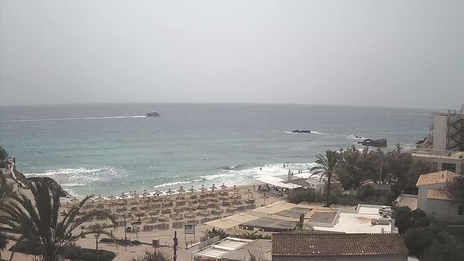 A hazy, overcast day reveals a beach with numerous umbrellas, a small boat on the ocean, and buildings overlooking the sandy shore.
