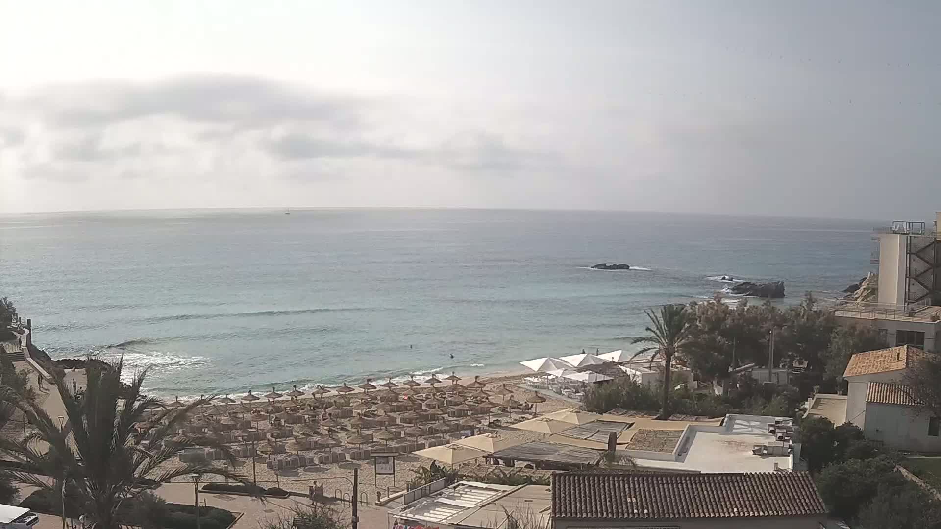 An overhead view shows a sandy beach with numerous umbrellas and beach chairs, a calm sea with small waves under a mostly cloudy sky, and buildings and palm trees situated on a coastal cliff.