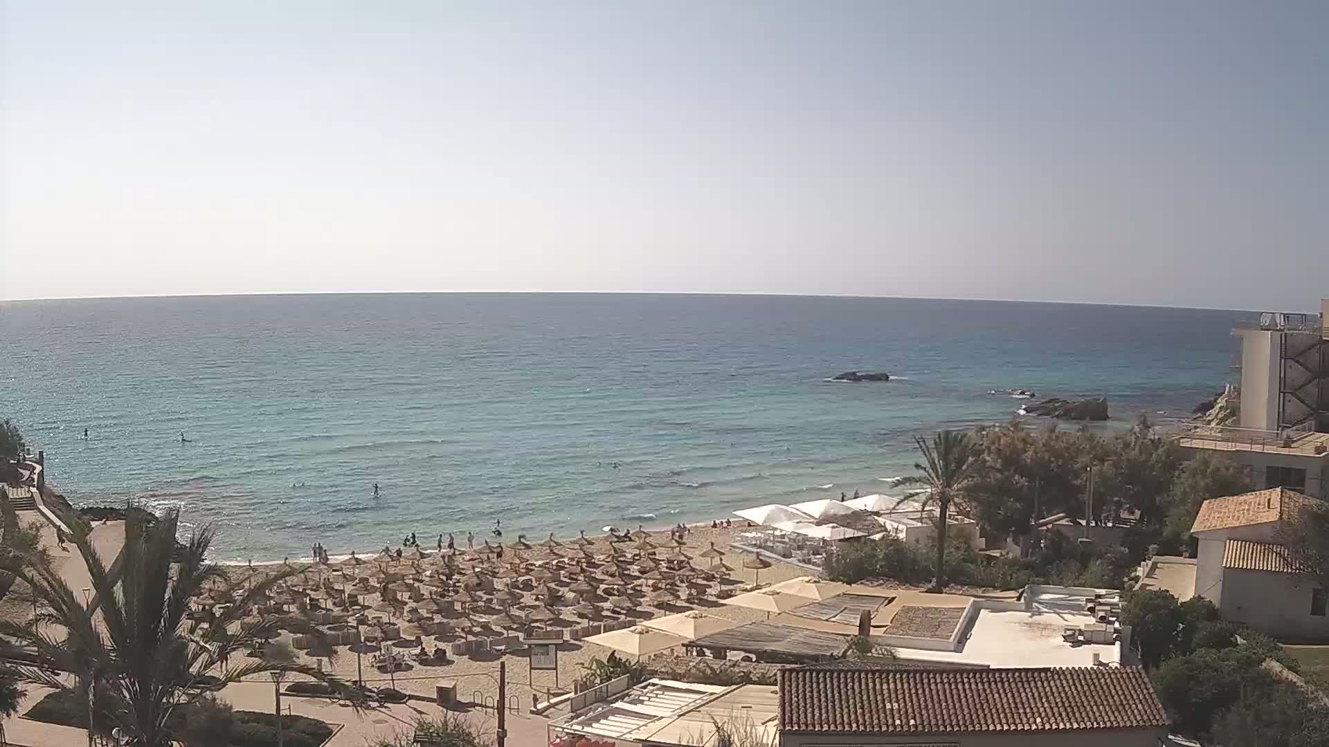 A sunny day at a beach with calm, light-blue water, numerous beach umbrellas, and several buildings overlooking the shore.