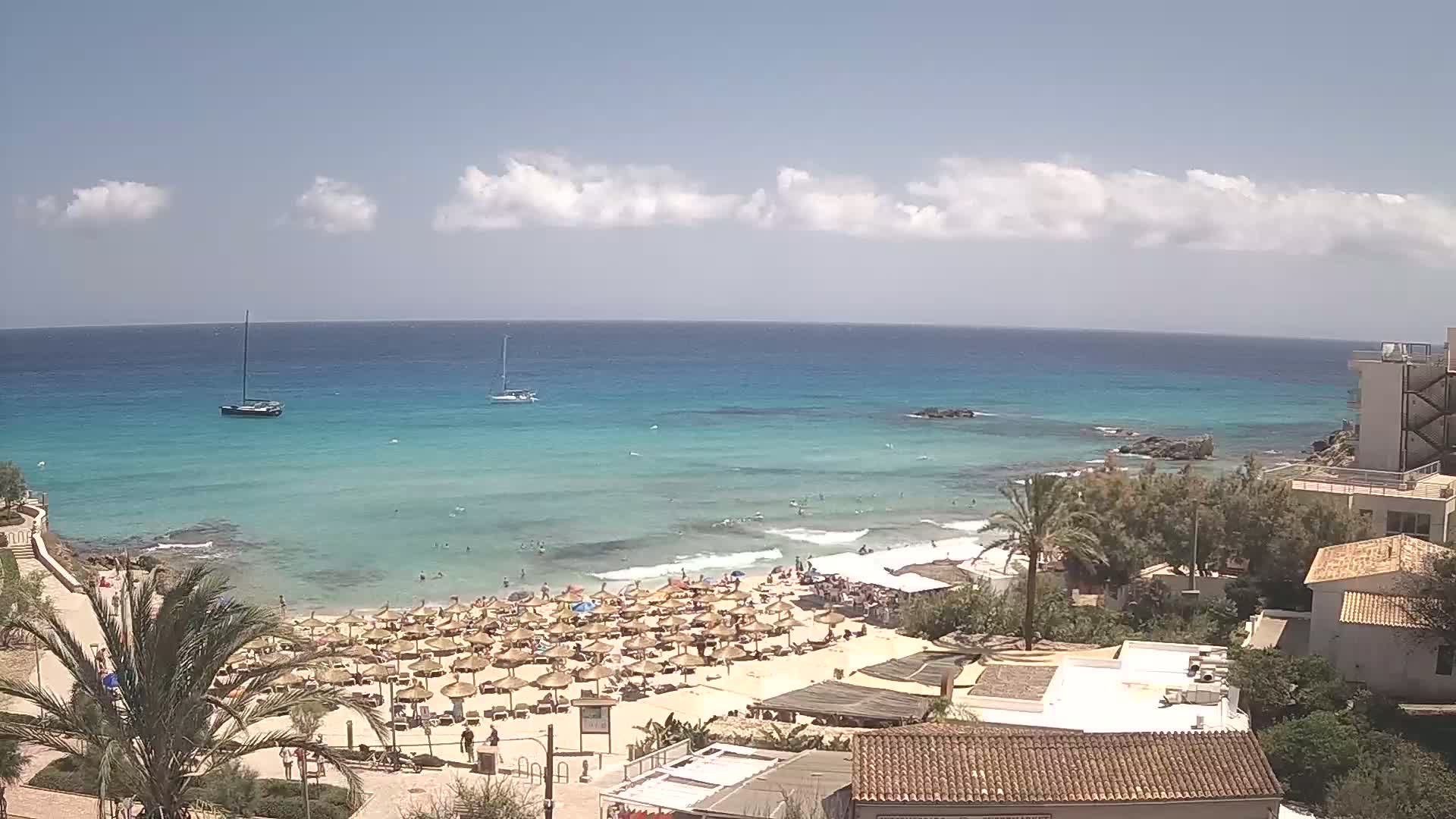 A sunny day at a beach with clear, turquoise water, numerous beach umbrellas, and several buildings overlooking the scene.