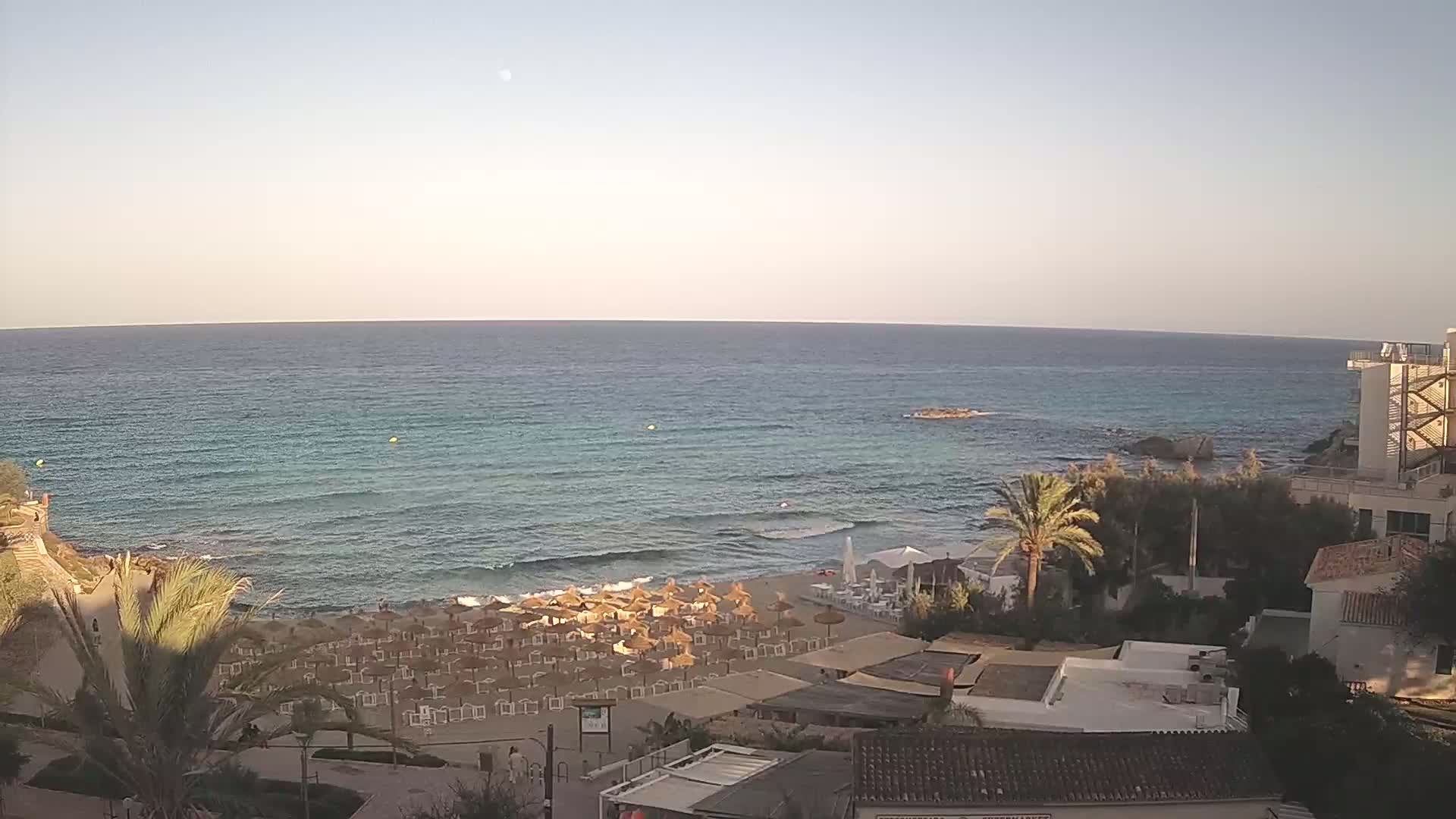 A sandy beach with numerous thatched umbrellas and beach chairs lines a calm, turquoise ocean under a clear, pale sky.