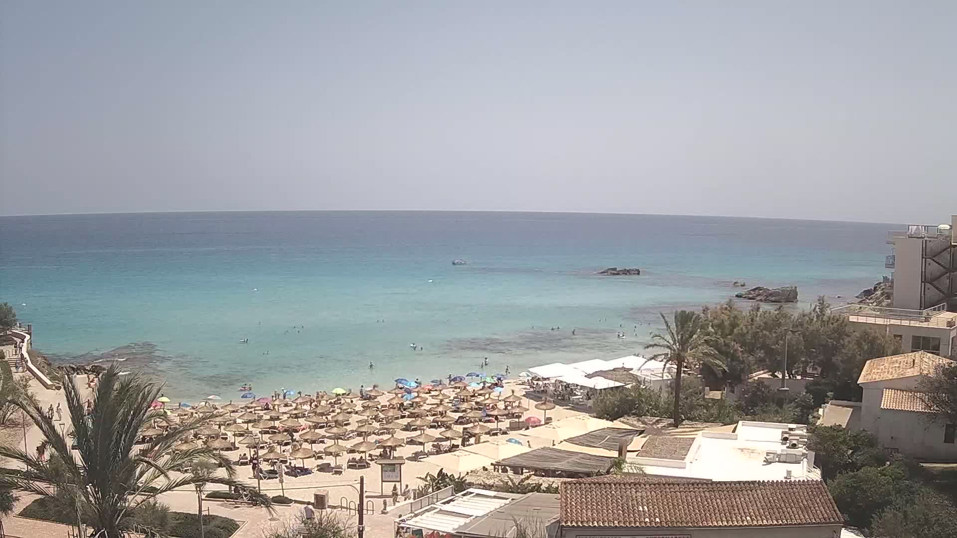 A sandy beach with numerous straw umbrellas and sunbathers is situated next to a calm, clear turquoise sea on a sunny day.
