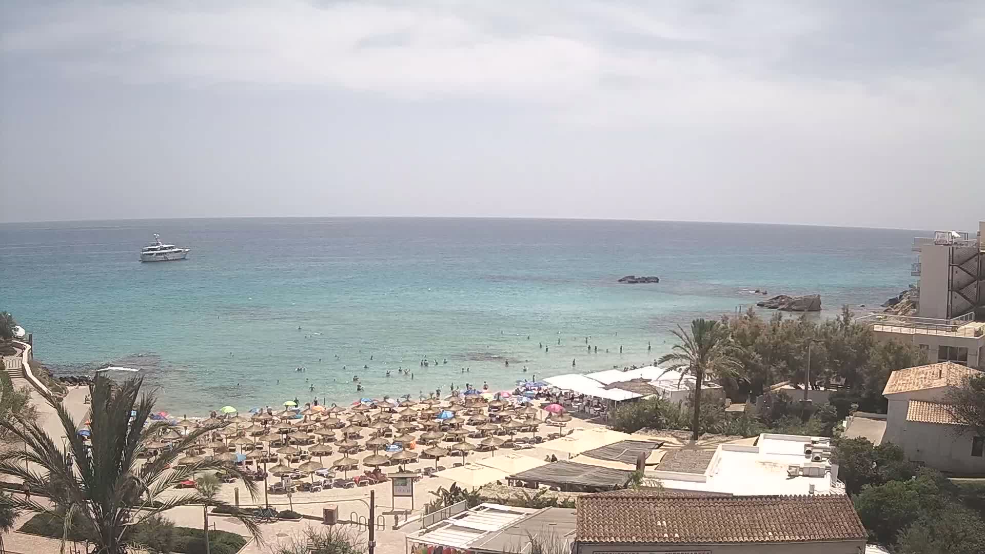 A crowded beach with numerous umbrellas and sunbathers is situated next to a calm, clear turquoise ocean under a partly cloudy sky, with a boat visible in the distance and buildings along the coast.