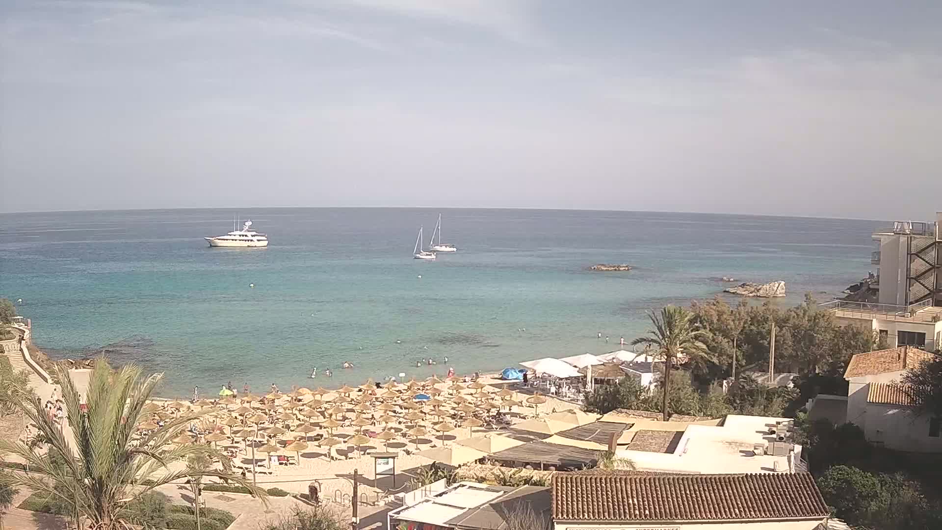 A sandy beach with numerous straw umbrellas and sunbathers is nestled between a calm, turquoise sea with boats and buildings overlooking the shore under a mostly clear sky.