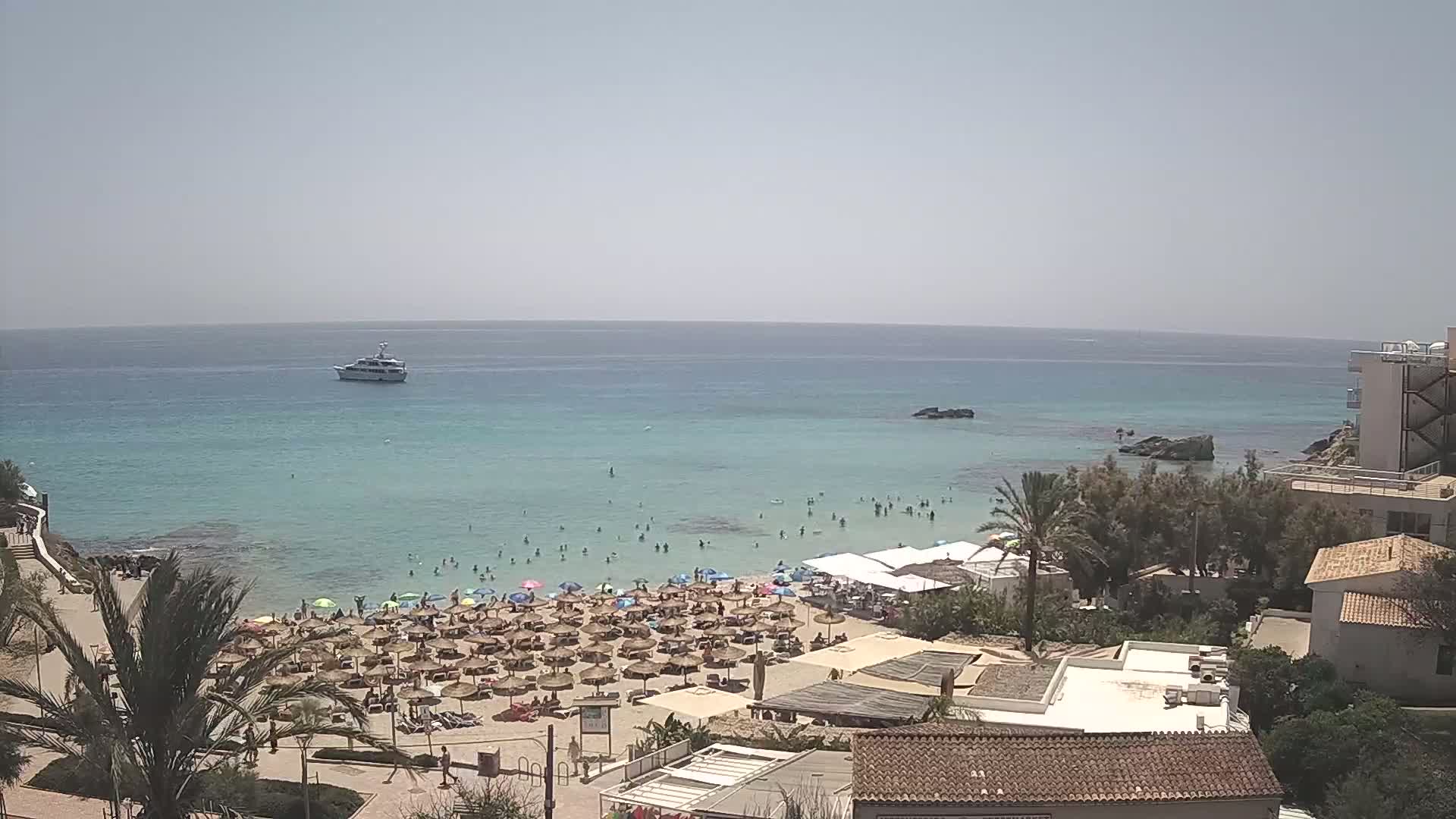 A sunny day at a beach with clear turquoise water, many people swimming and sunbathing under numerous umbrellas, and a boat visible in the distance.