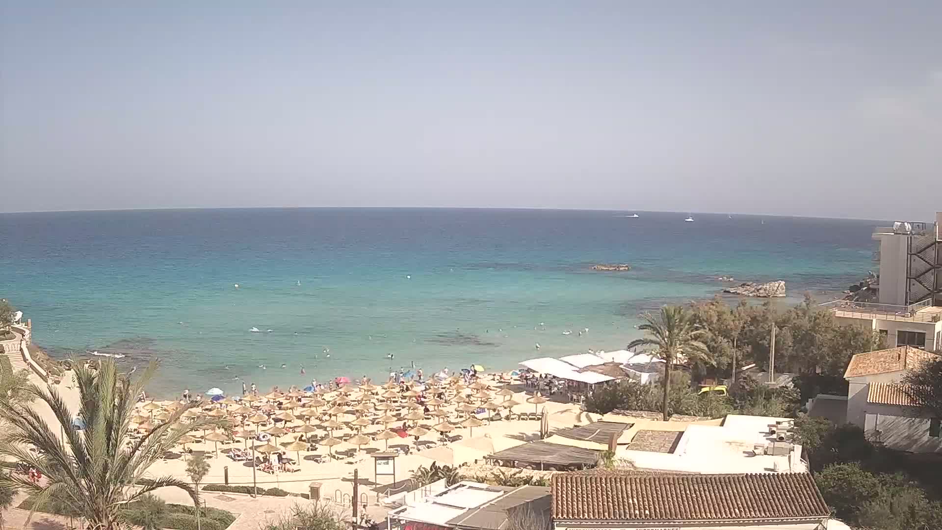 A sandy beach with numerous straw umbrellas and sunbathers overlooks a calm, clear turquoise ocean under a bright, sunny sky.