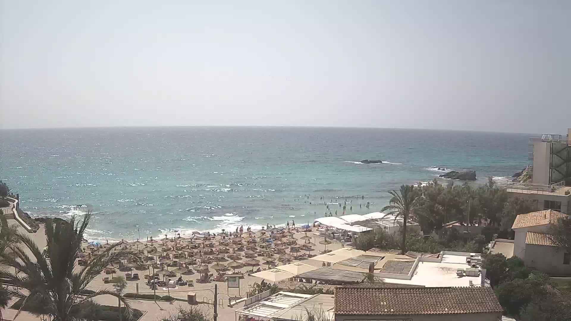 A sandy beach with numerous umbrellas and people is situated next to a turquoise ocean with small waves under a bright, mostly clear sky.
