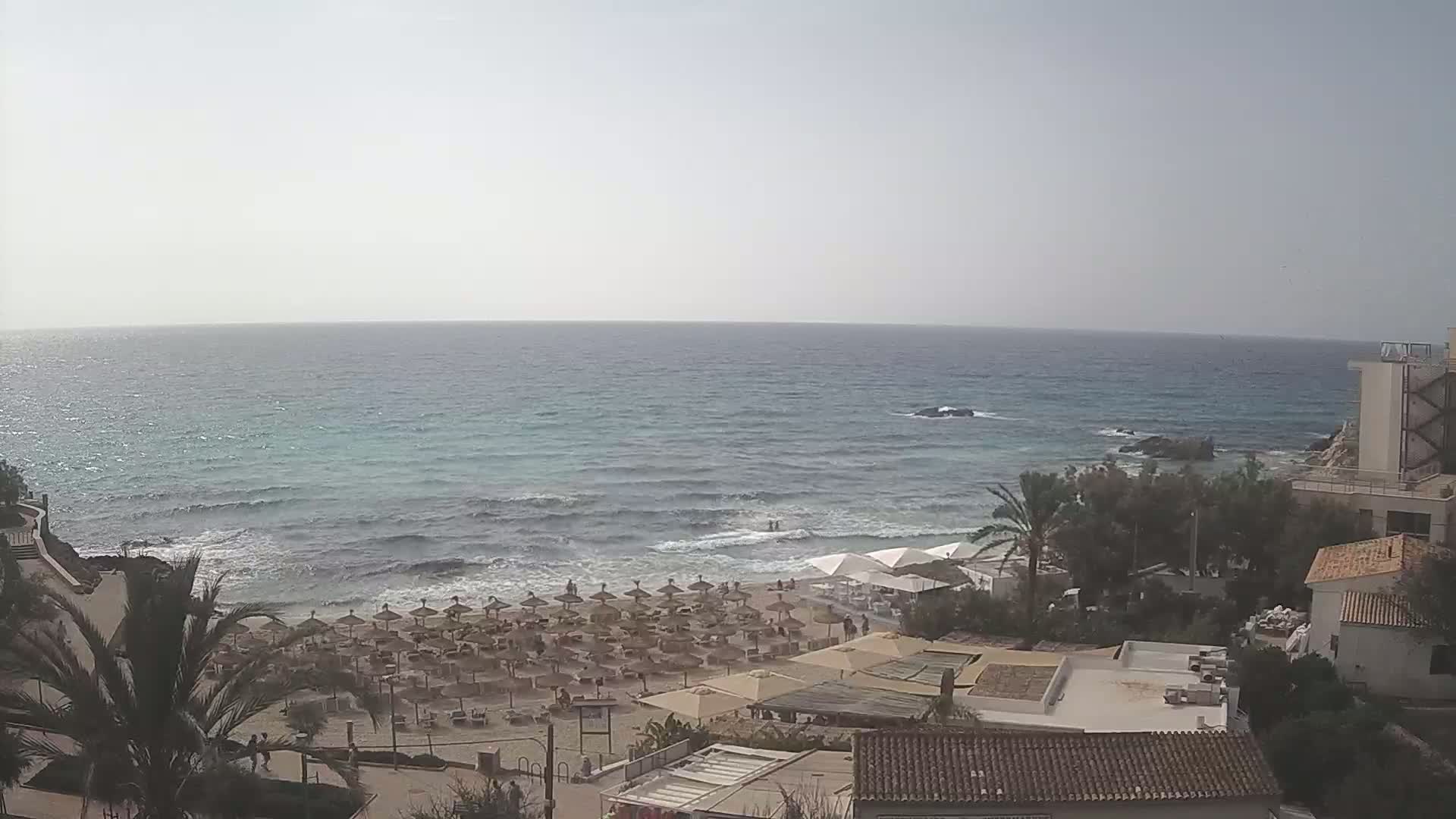 A beach with numerous umbrellas and lounge chairs is visible from an elevated viewpoint overlooking a calm sea with small waves under a hazy sky.