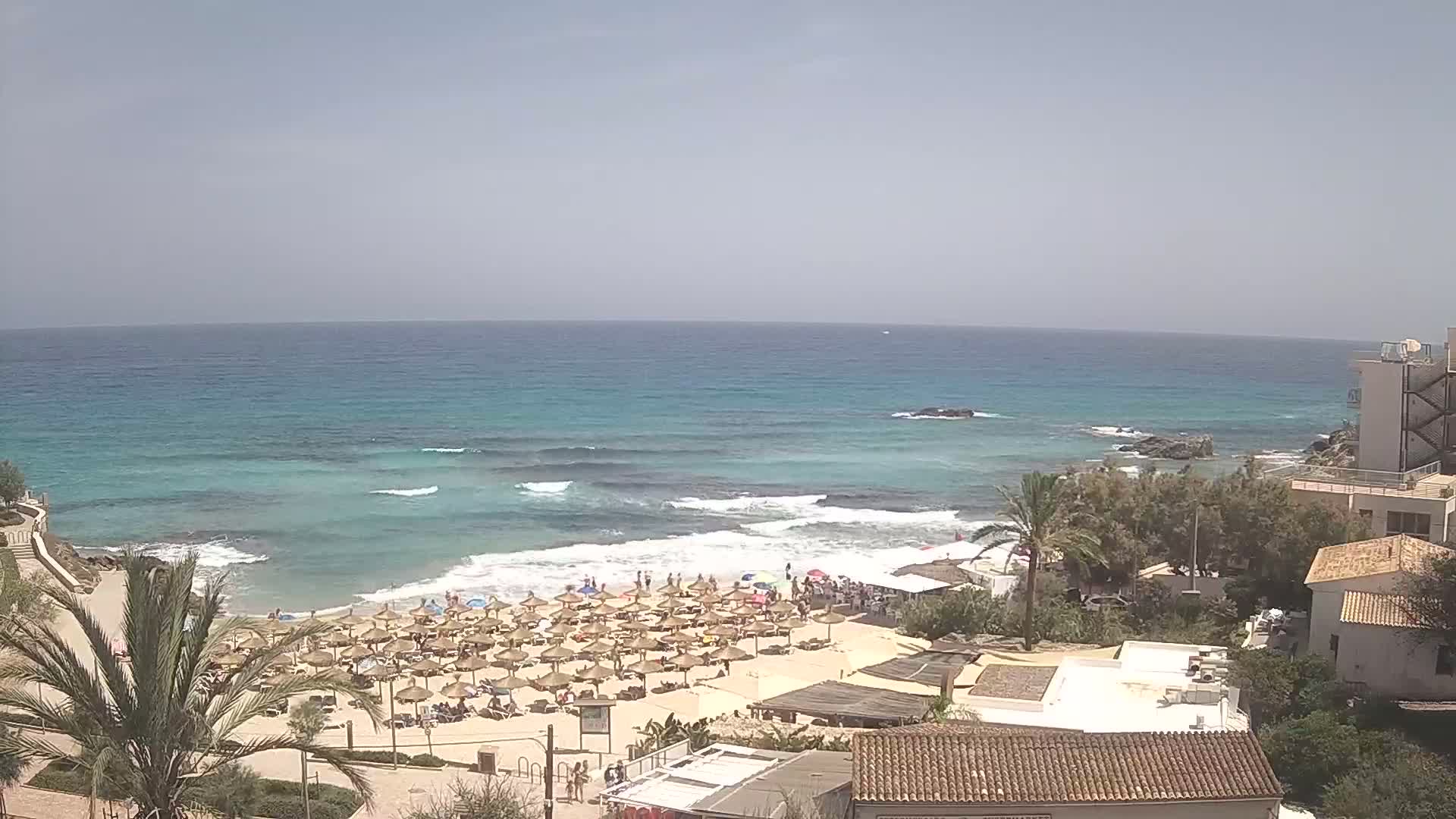 A sunny day at a beach with light waves, numerous umbrellas on the sand, and buildings overlooking the scene.