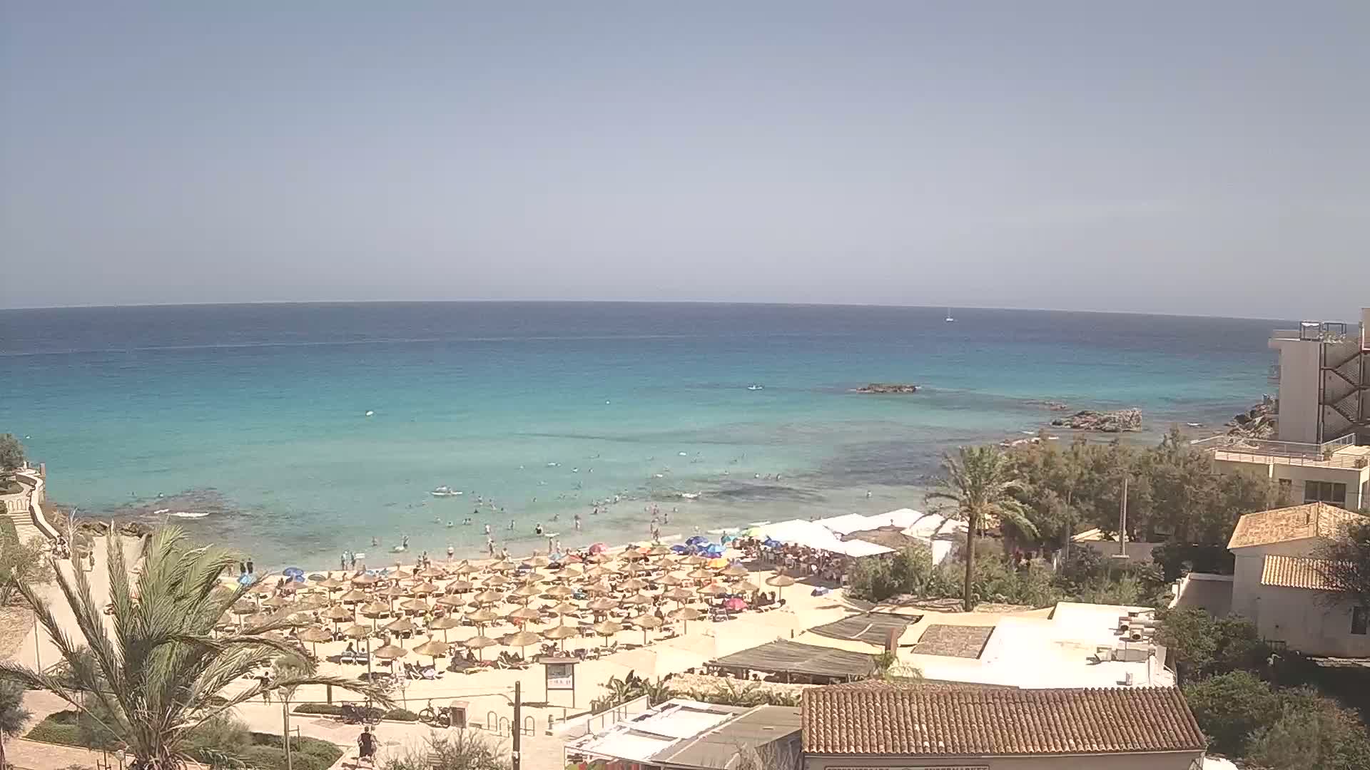 A sunny day at a beach shows many straw umbrellas and sunbathers on the sand, with clear turquoise water extending to the horizon.