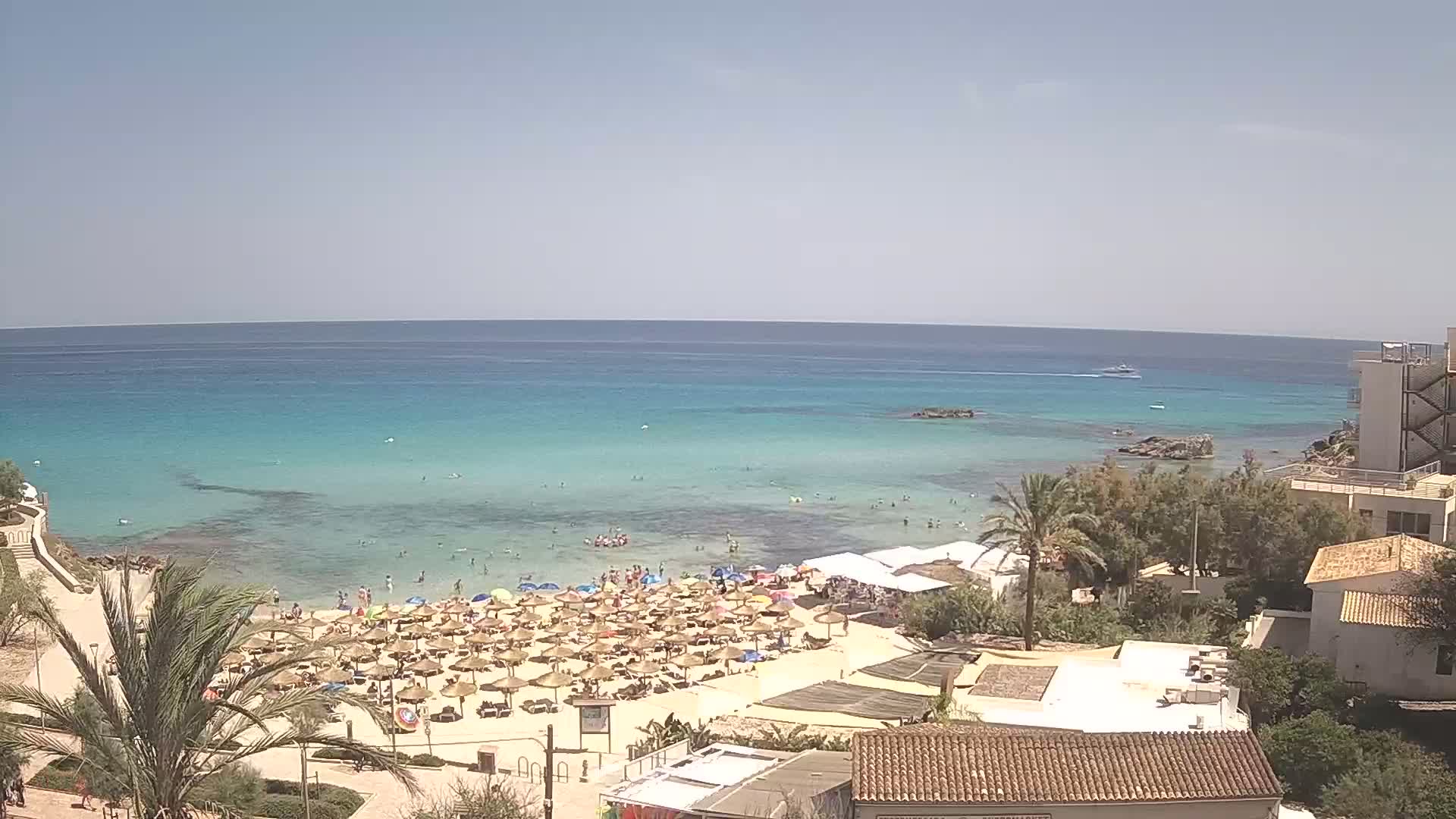 A sunny day at a beach with clear turquoise water, numerous beach umbrellas, and several buildings are visible overlooking the coastline.