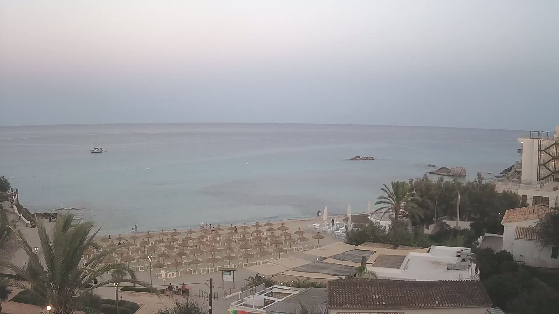 An overcast day reveals a beach with numerous straw umbrellas and lounge chairs, a calm sea with a boat, and buildings situated on a hillside overlooking the water.