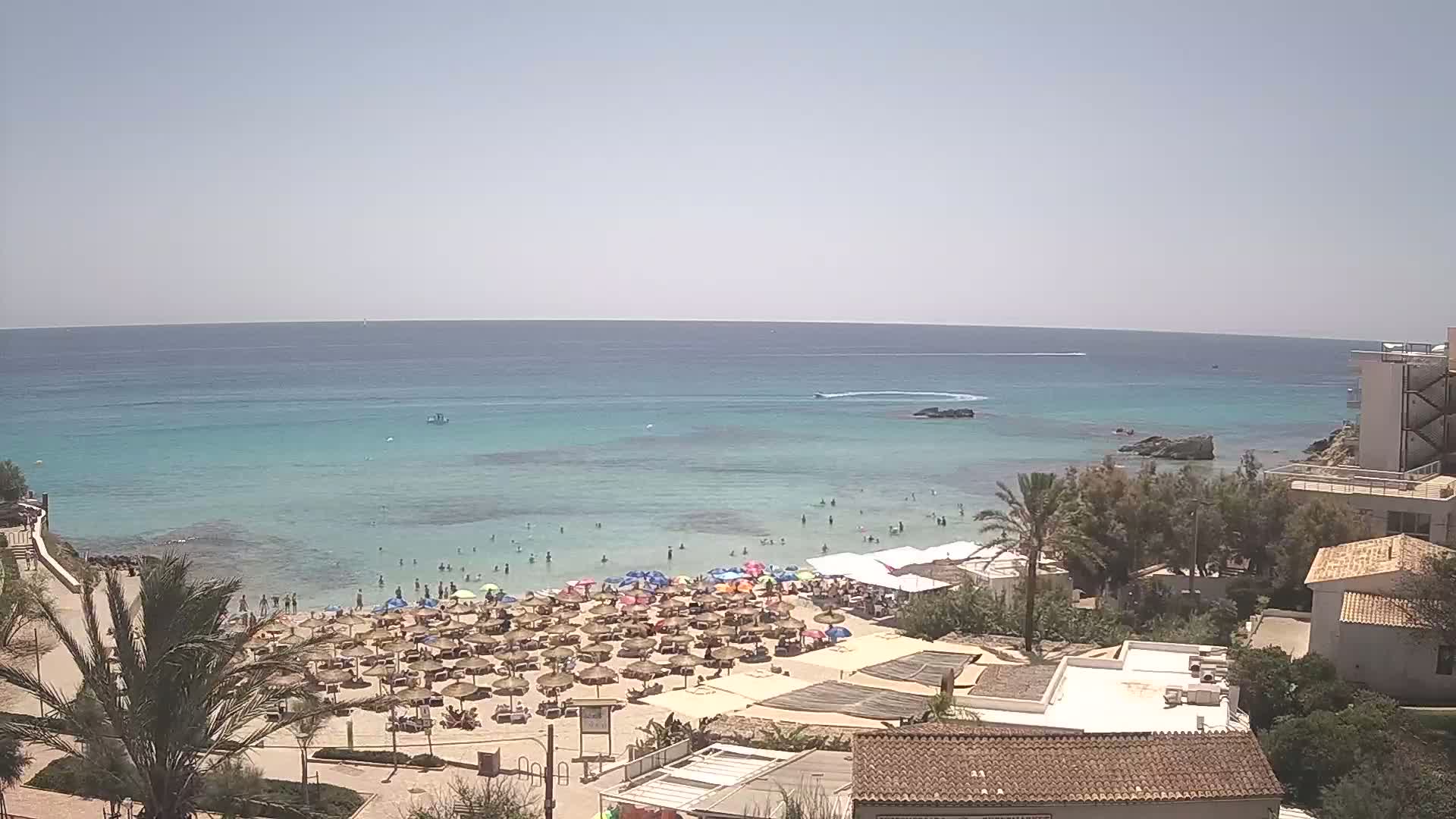 A crowded beach with numerous umbrellas and sunbathers stretches along a calm, turquoise sea under a clear sky.