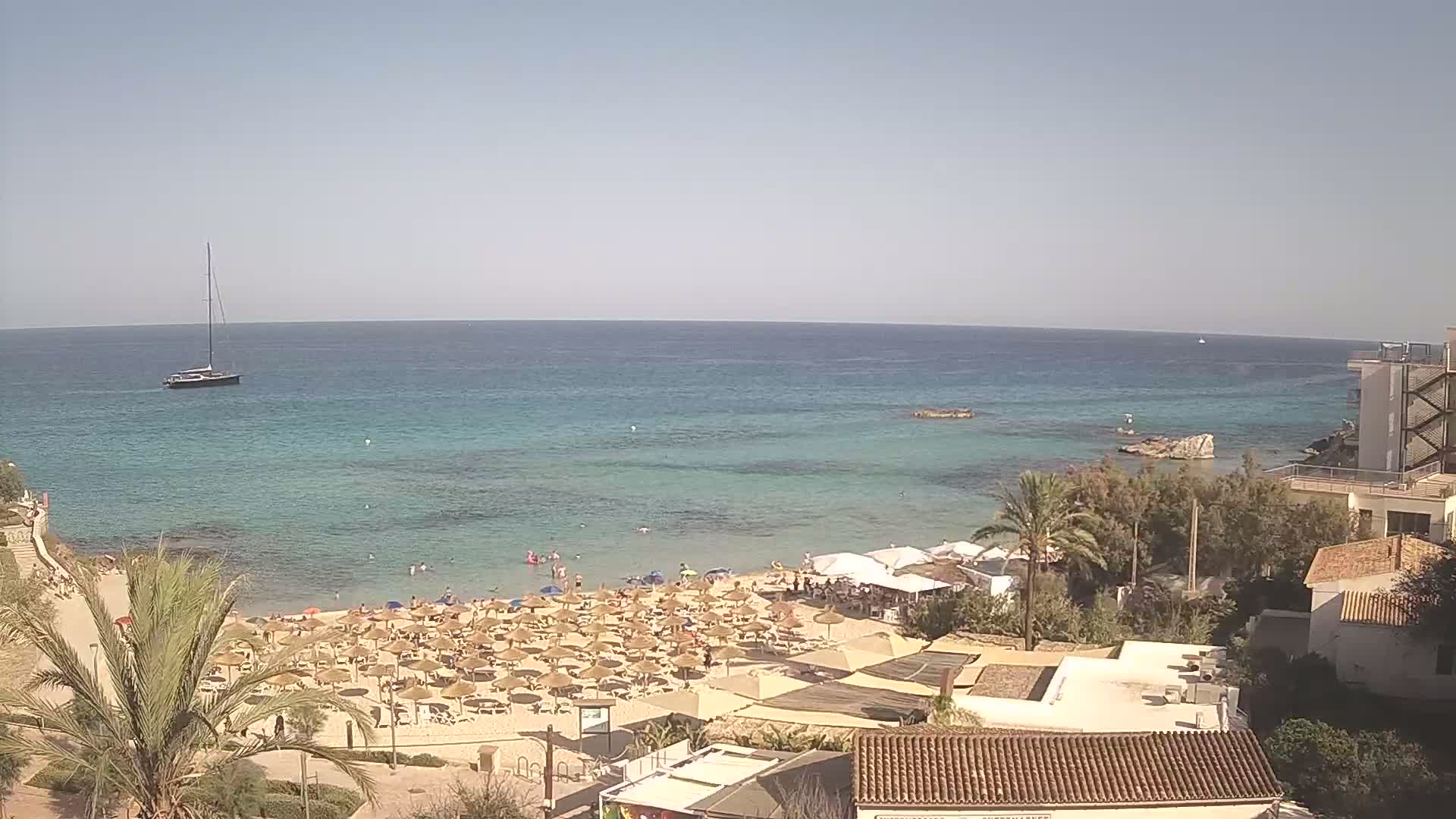 A sandy beach with numerous umbrellas and sunbathers is situated next to a calm, clear turquoise sea under a bright, sunny sky, with a sailboat visible in the distance and buildings overlooking the scene.