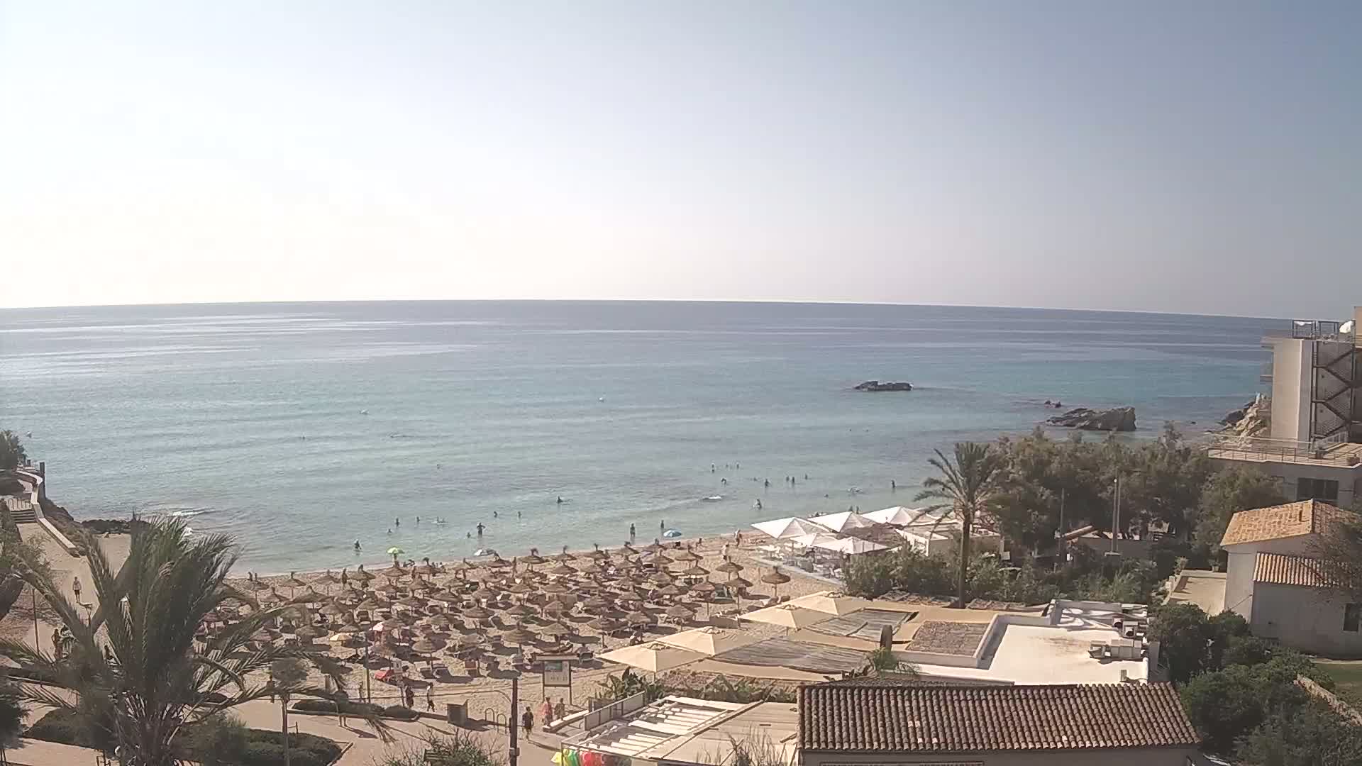 A sunny day at a beach with calm, light-blue water, numerous umbrellas and sunbathers on the sand, and buildings and vegetation on the adjacent land.