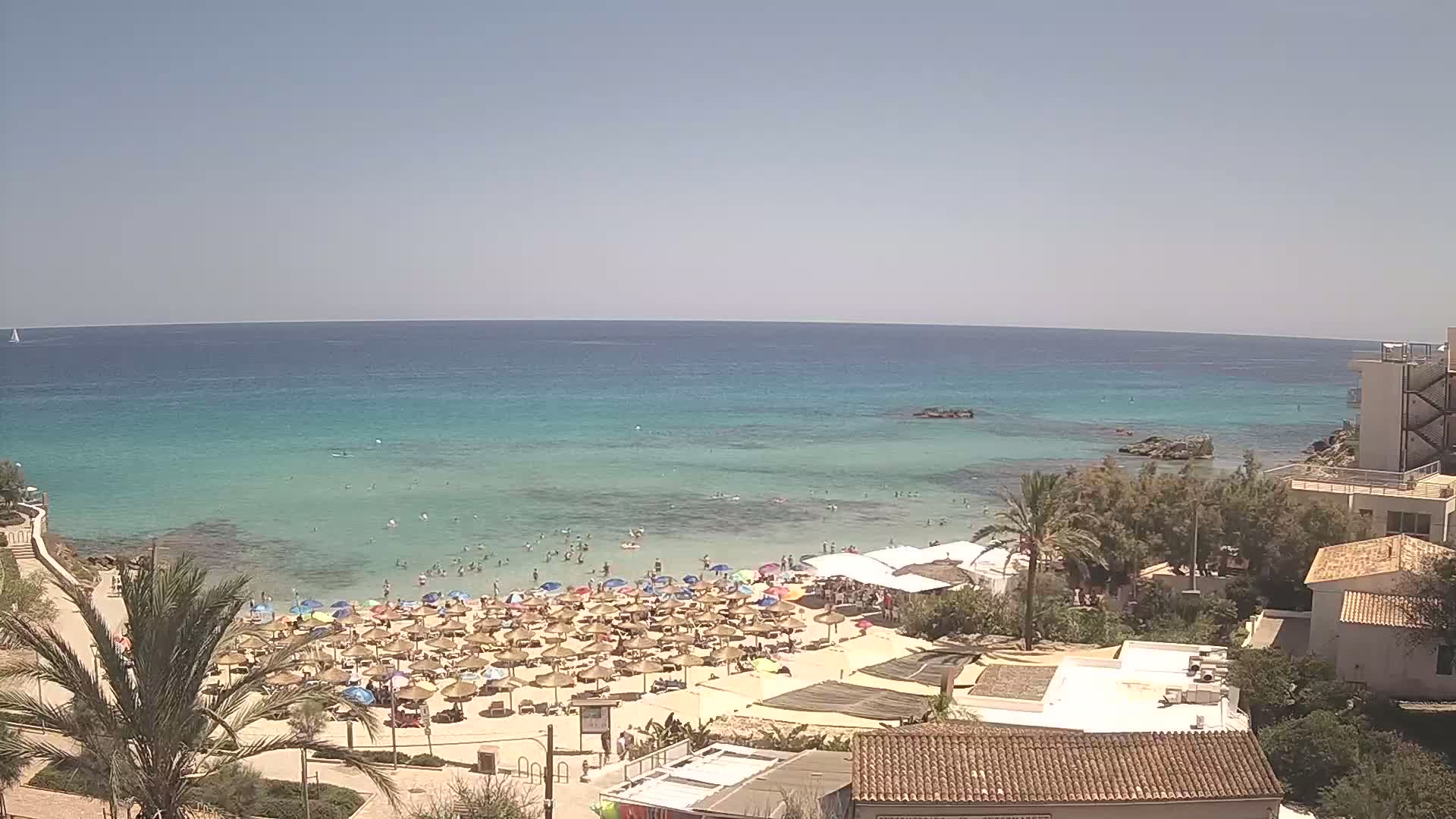 A sandy beach with numerous umbrellas and bathers is situated next to a calm, turquoise ocean under a clear blue sky.
