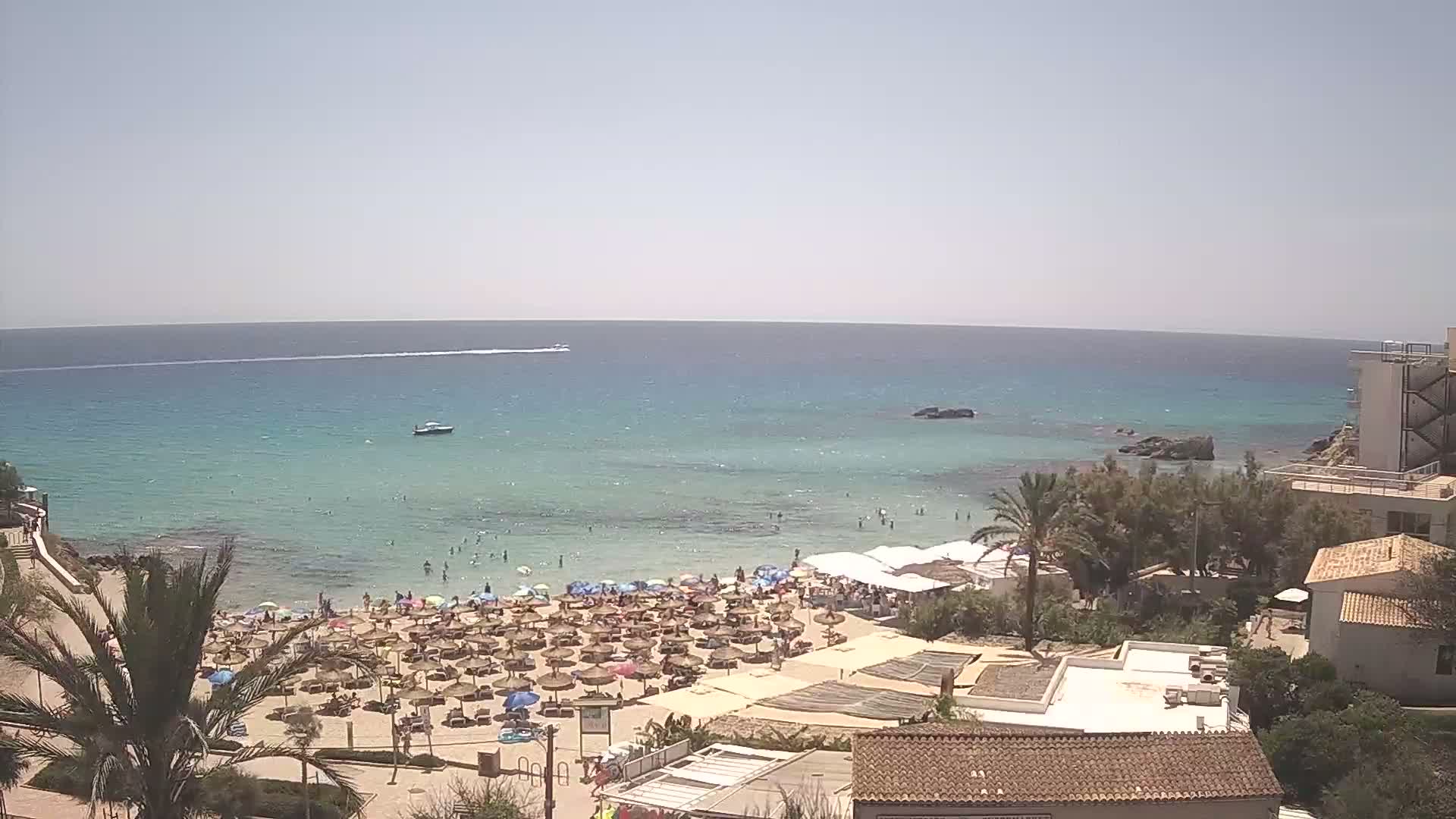 A sunny day at a beach with clear, shallow turquoise water, numerous beach umbrellas and sunbathers, and buildings situated on a hillside overlooking the scene.
