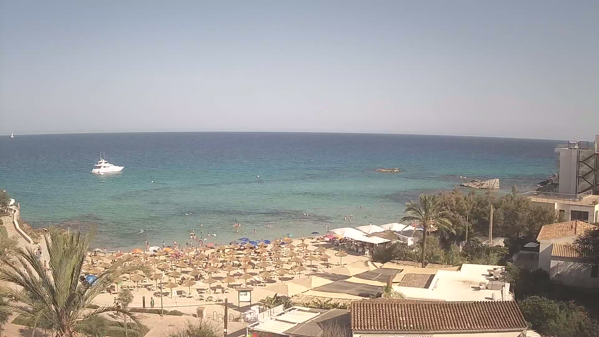 A beach with numerous umbrellas and sunbathers is visible alongside a calm, clear turquoise sea on a sunny day, with a boat in the distance and buildings and vegetation in the foreground.