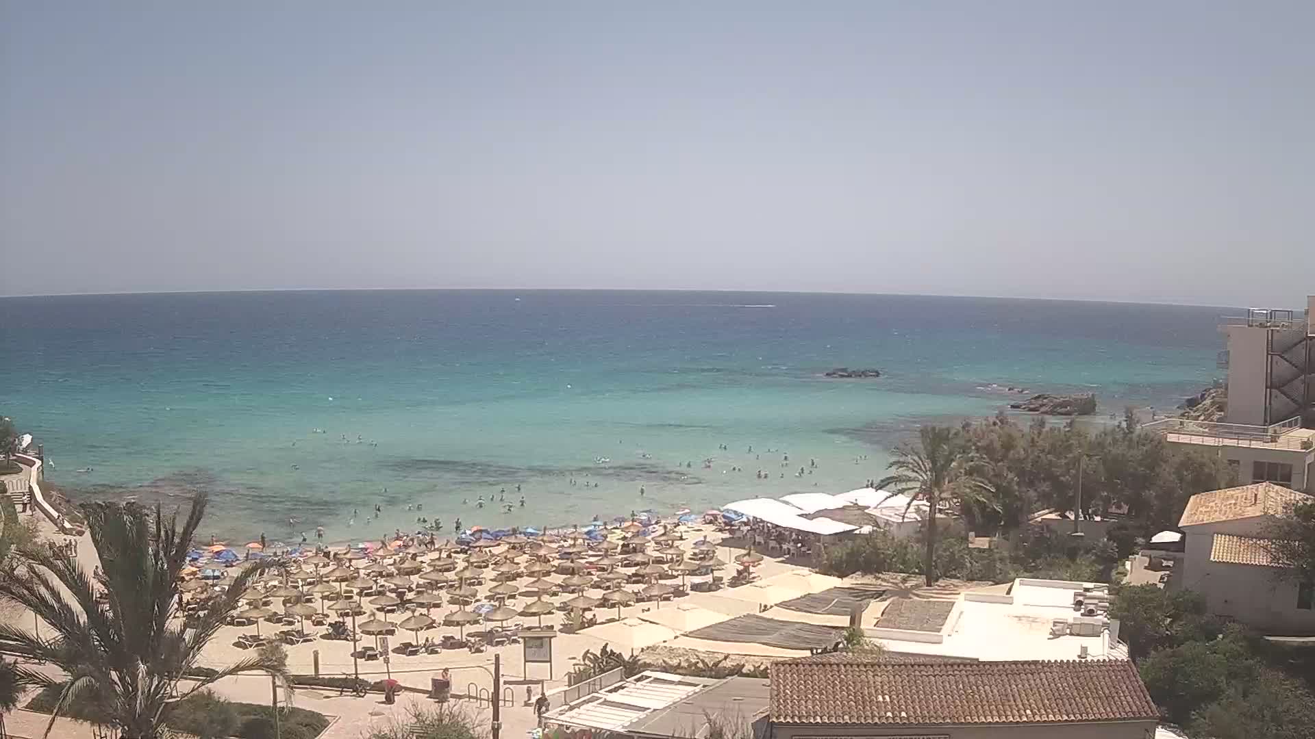 A sandy beach with numerous umbrellas and sunbathers is situated next to a calm, clear turquoise sea on a sunny day.