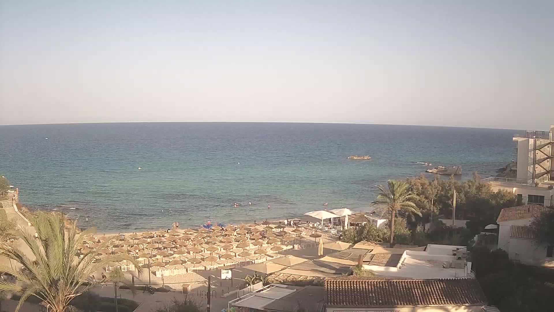A sandy beach with numerous umbrellas and a few people in the water is situated next to a calm, clear ocean under a clear sky.