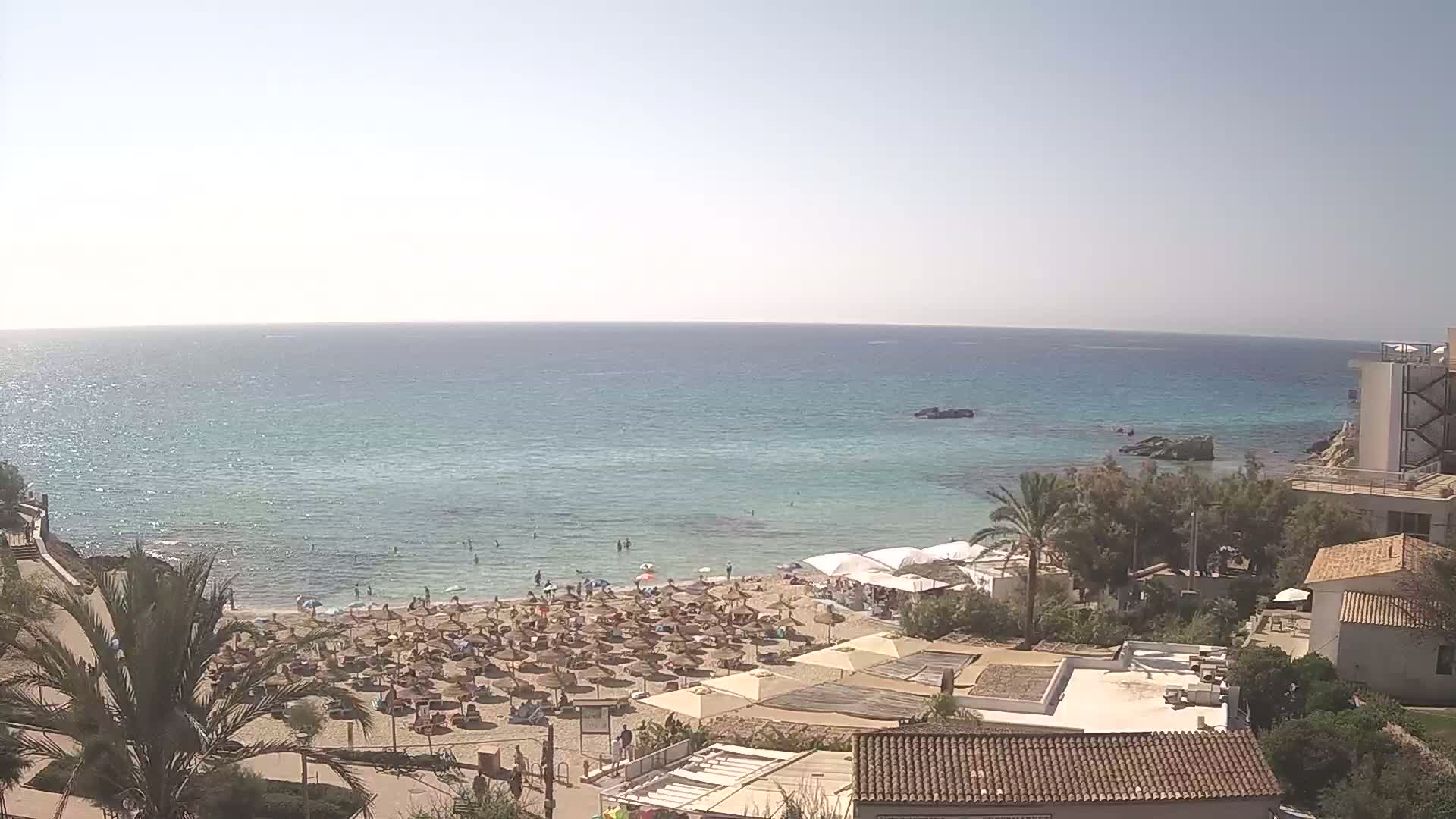 A sandy beach with numerous umbrellas and sunbathers is situated next to a calm, clear turquoise sea under a bright, sunny sky.