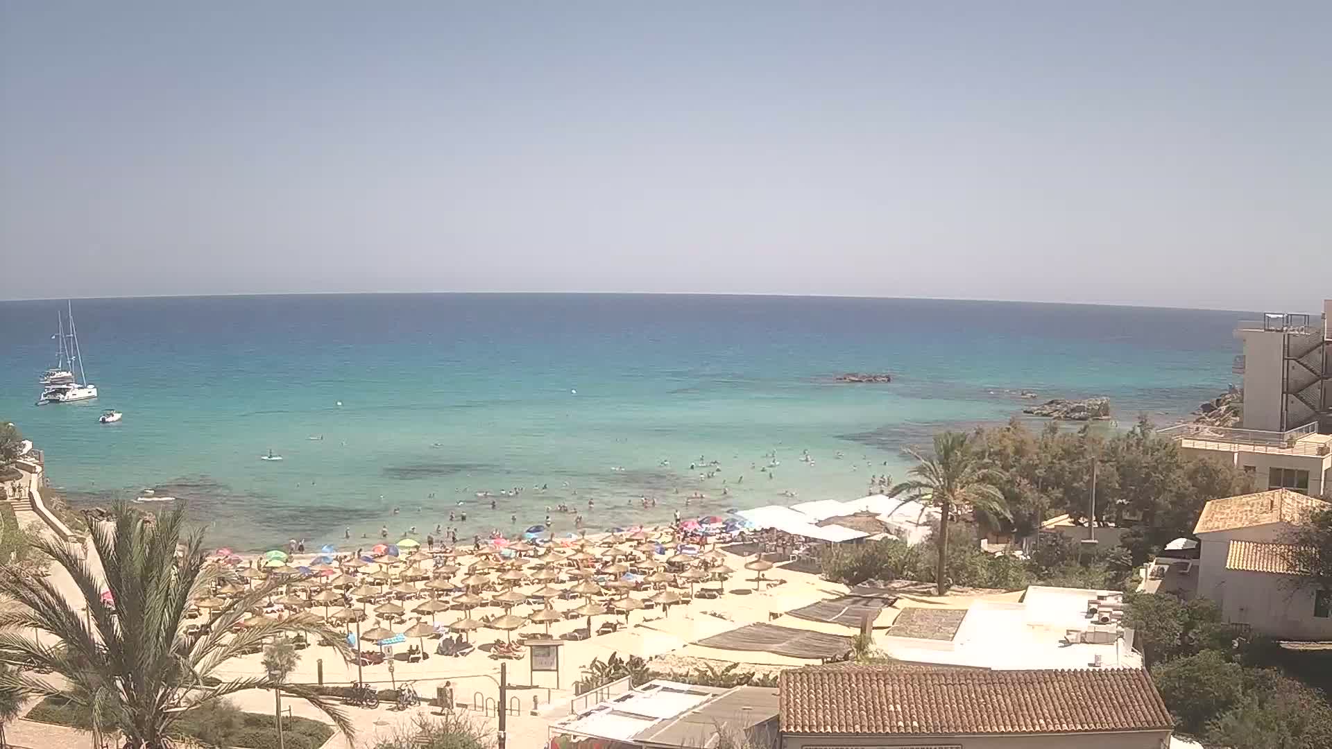A sunny day at a crowded beach with clear turquoise water, numerous beach umbrellas, and buildings overlooking the coast.