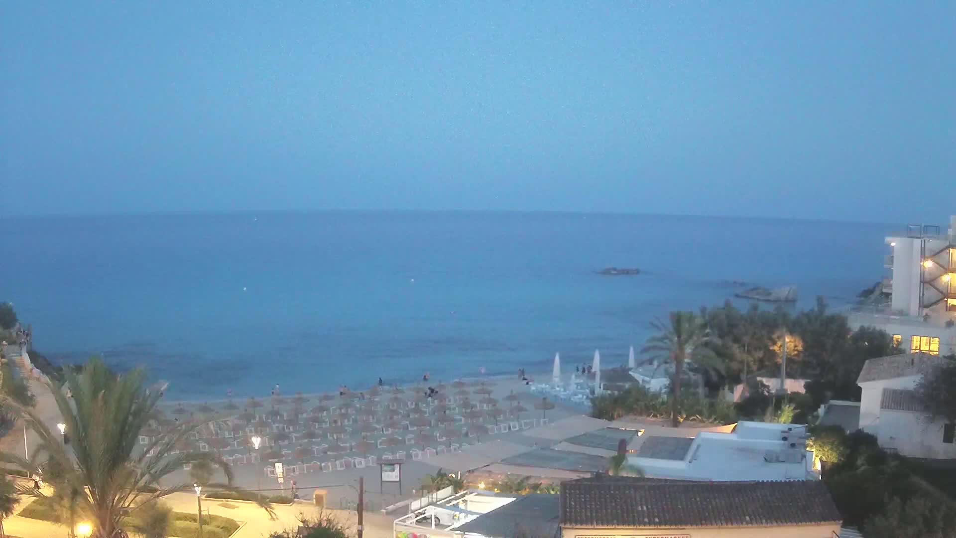 A beach with numerous lounge chairs and umbrellas sits below a collection of buildings at dusk, overlooking a calm, blue sea.