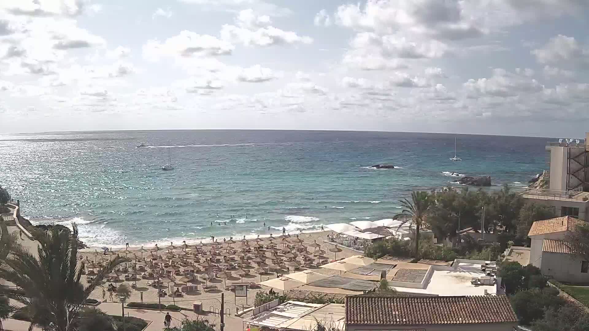 A partly cloudy day shows a sandy beach with numerous umbrellas and people, a calm ocean with a few boats, and buildings along the shoreline.