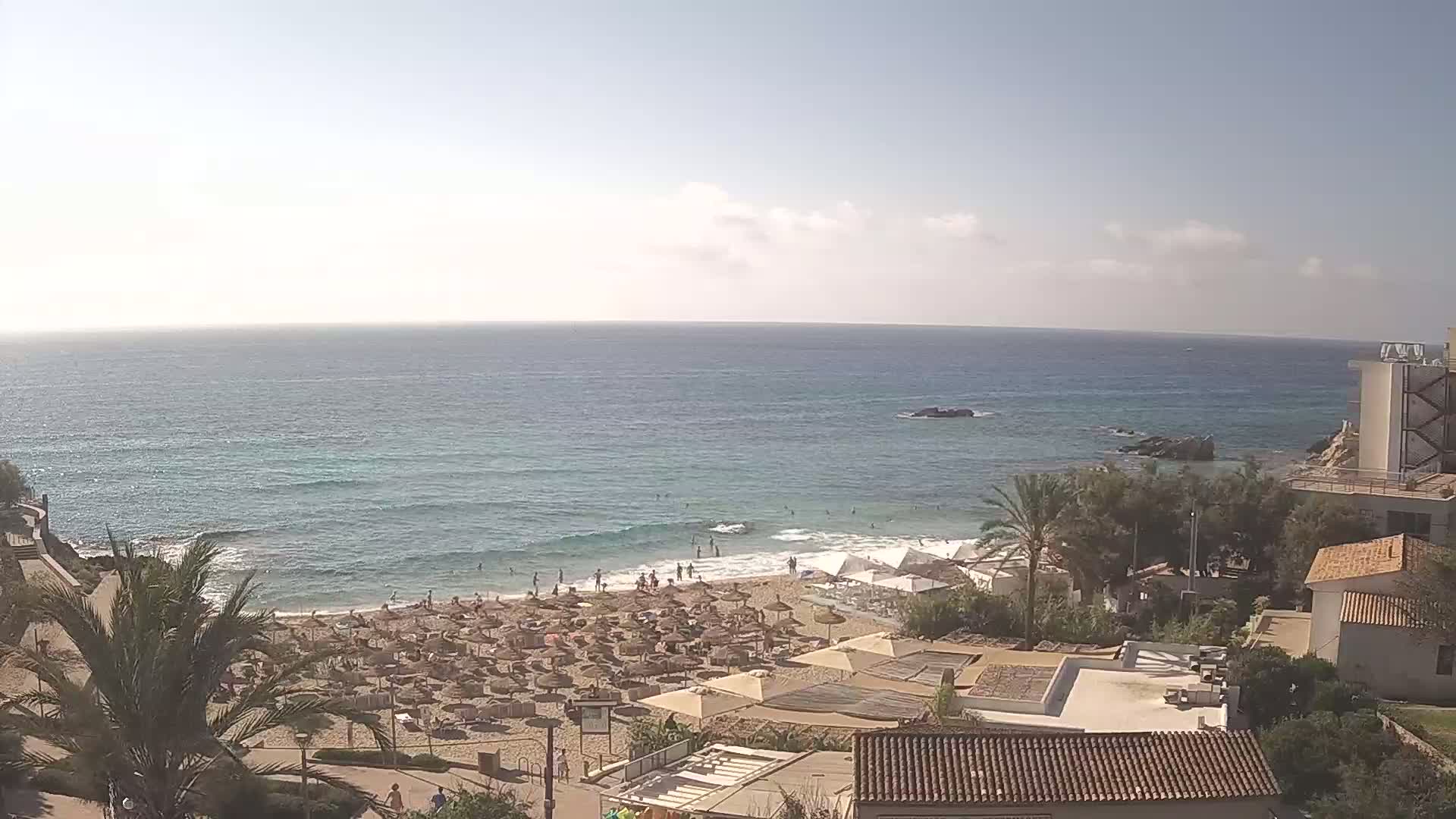 A sandy beach with numerous umbrellas and sunbathers is situated next to a calm, blue ocean under a partly sunny sky.