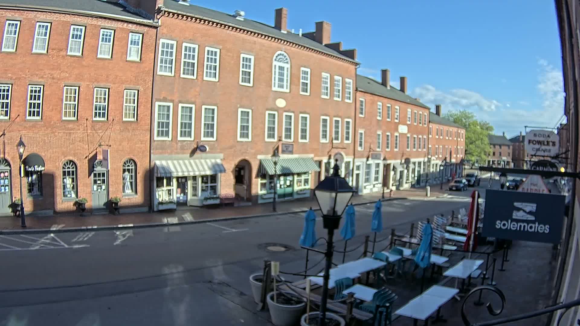 A sunny day on a brick-lined street with several shops and a cafe patio with blue umbrellas.