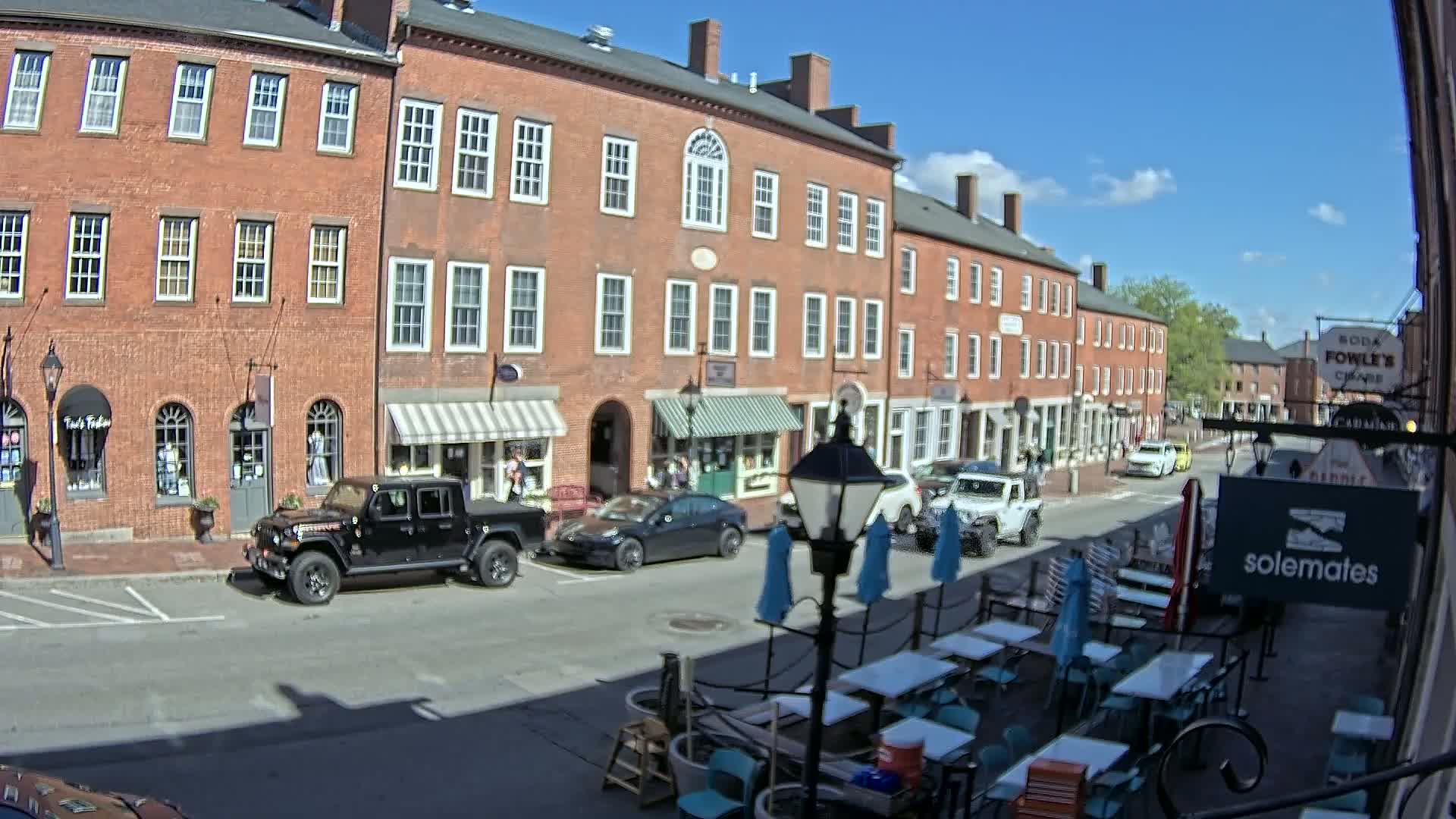 A sunny day reveals a brick street lined with shops and restaurants, featuring outdoor seating and parked cars.