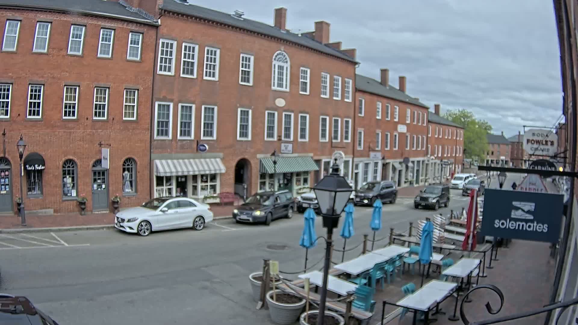 A street scene on an overcast day shows brick buildings with shops and outdoor seating, several cars parked and driving, and a few pedestrians.