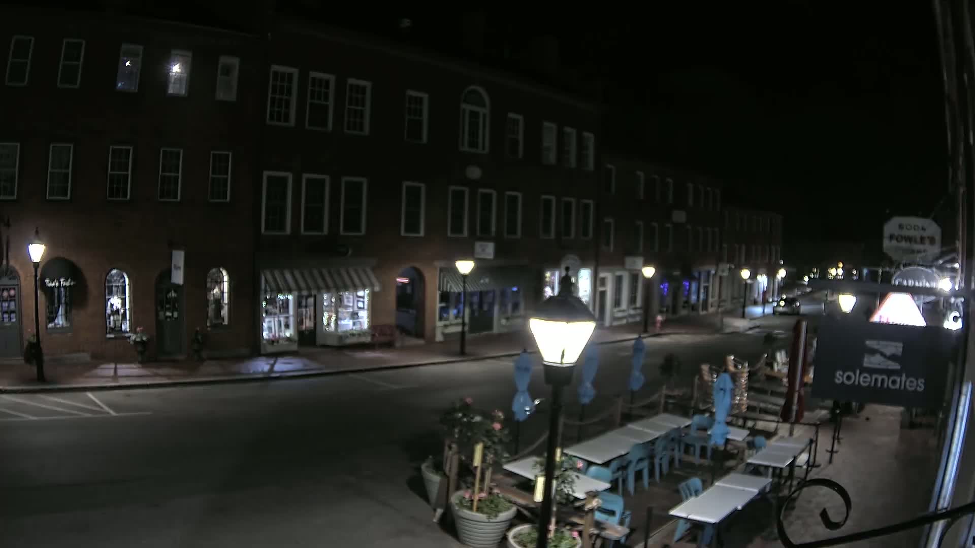 A nighttime view of a brick street with shops and a restaurant patio, lit by streetlamps.