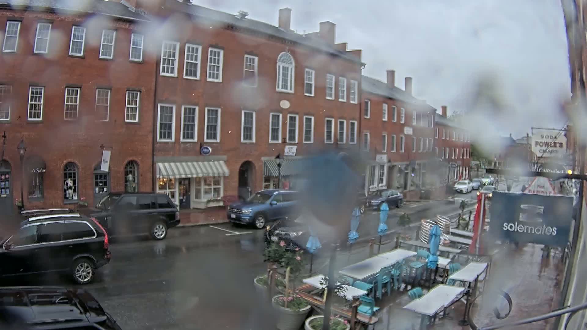 A rainy view from inside a building shows a brick street with parked cars, outdoor seating, and several shops.