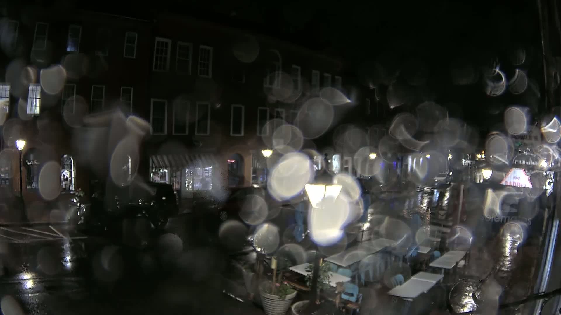 A rain-streaked window shows a nighttime view of a street with buildings, streetlights, and outdoor seating at a restaurant.