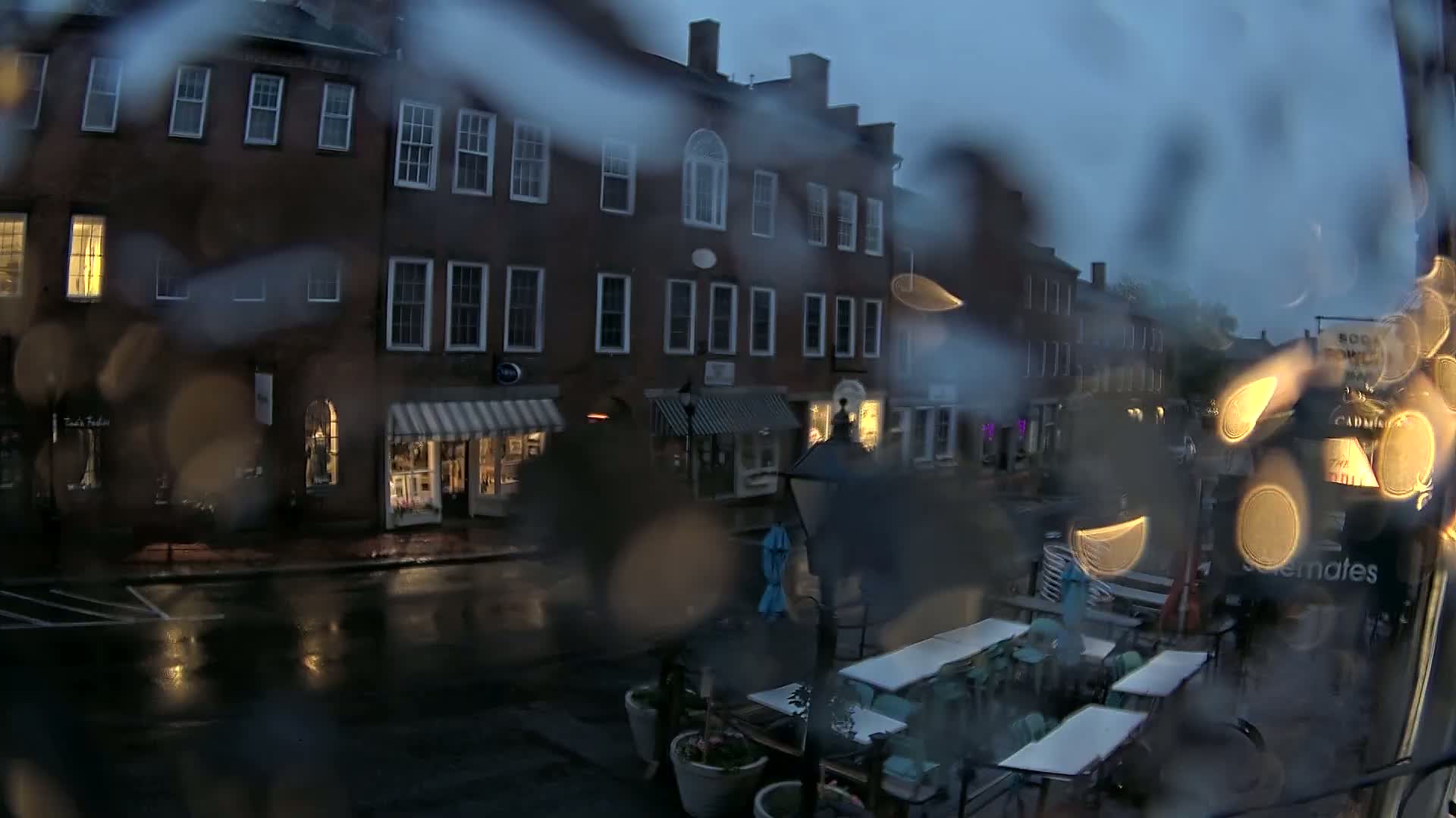 A rain-streaked window view shows a dimly lit street with brick buildings and outdoor seating at a restaurant under a dark, overcast sky.