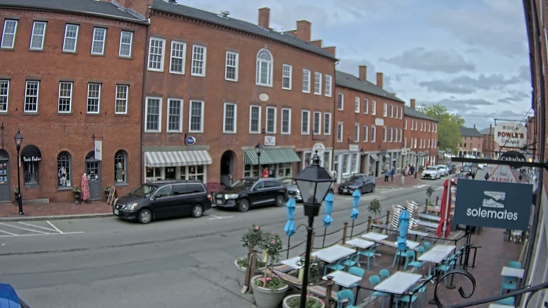 A street scene on a partly cloudy day shows a row of brick buildings with shops and awnings, parked cars, pedestrians, and an outdoor patio seating area with teal chairs and tables.