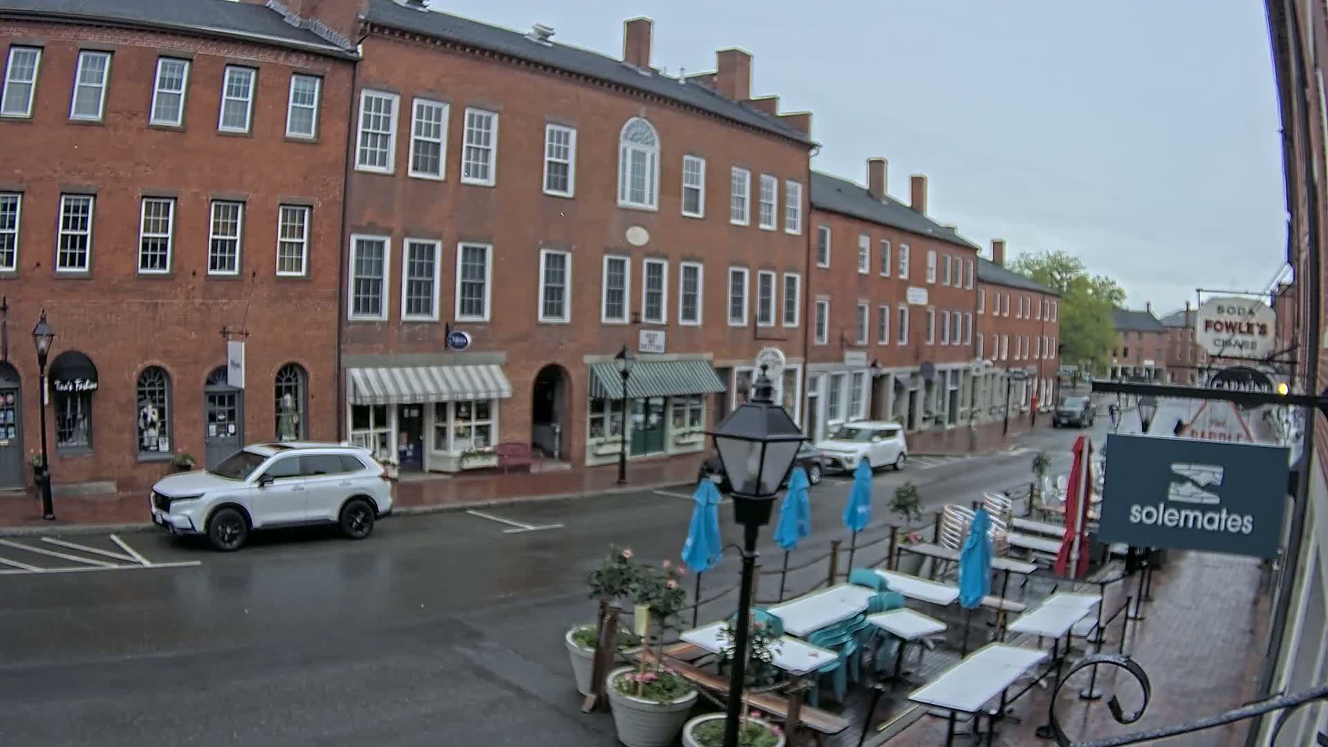 A wet street scene shows a row of brick buildings with awnings and outdoor seating, and a white SUV is parked along the curb.
