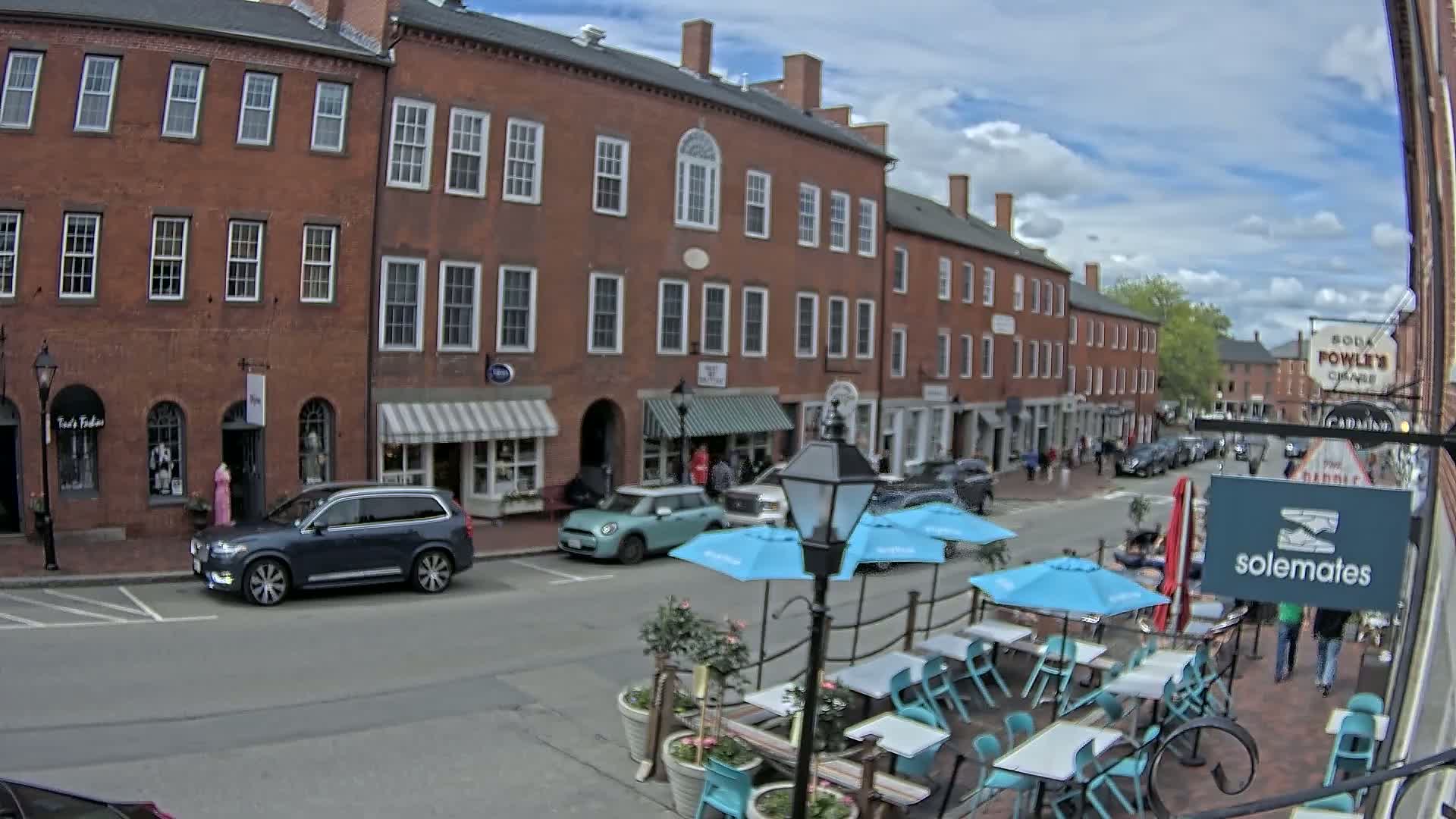 A partly sunny day reveals a brick street lined with shops and a patio dining area with light blue umbrellas.