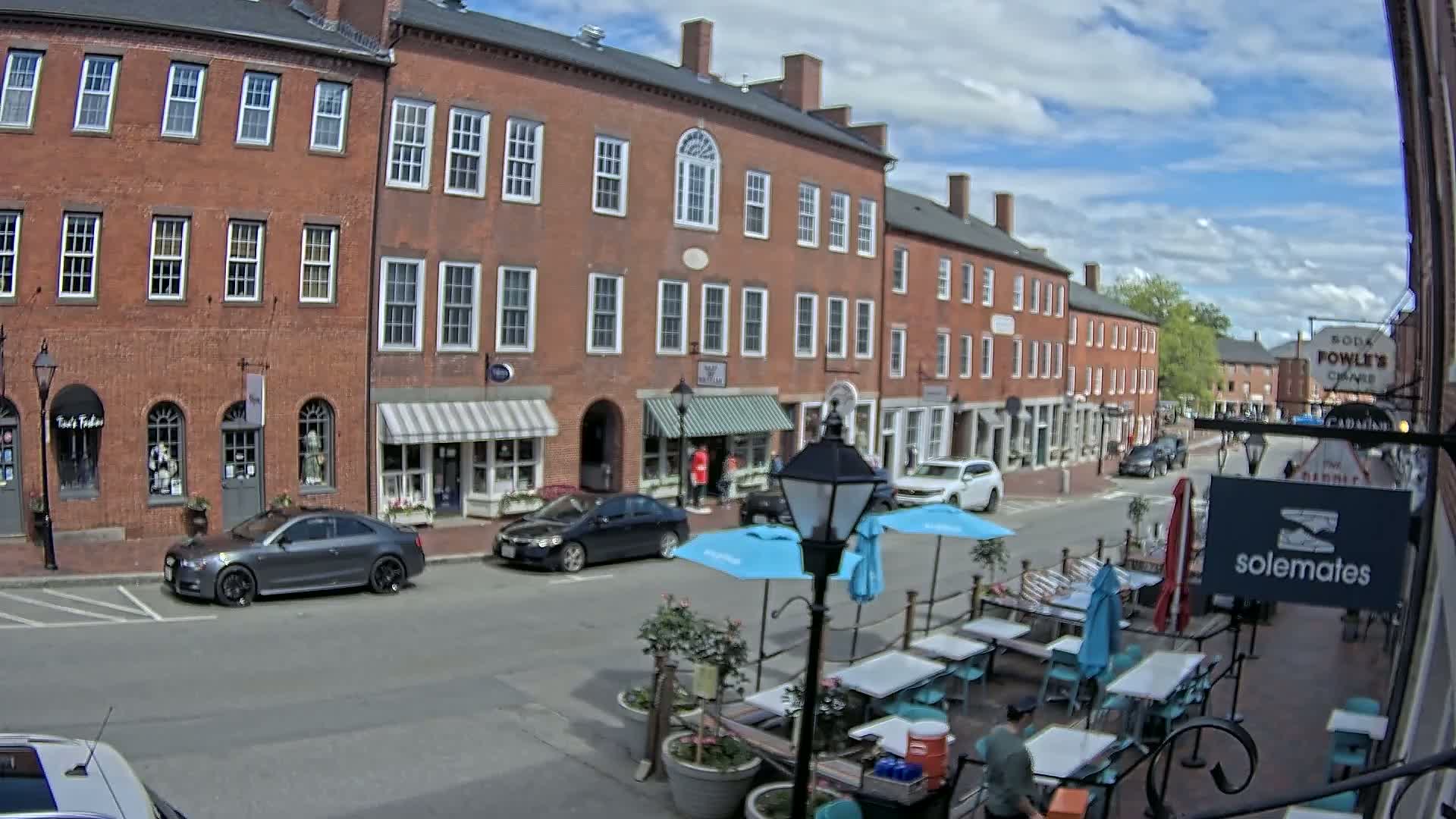 A partly sunny day reveals a brick street lined with shops and a patio dining area with light blue umbrellas.